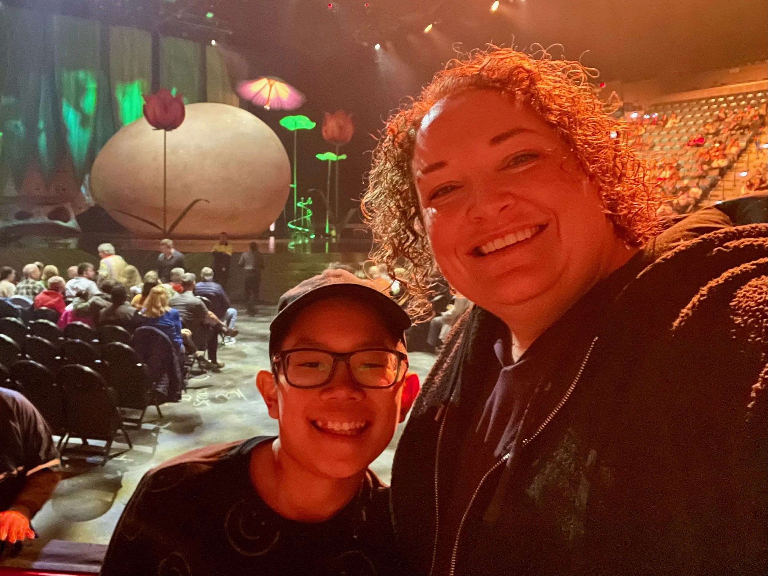 Two smiling individuals, a young person with glasses and an older woman, taking a selfie at a theater with a stage decorated with large, colorful mushroom and flower props. Audience can be seen seated in the background.