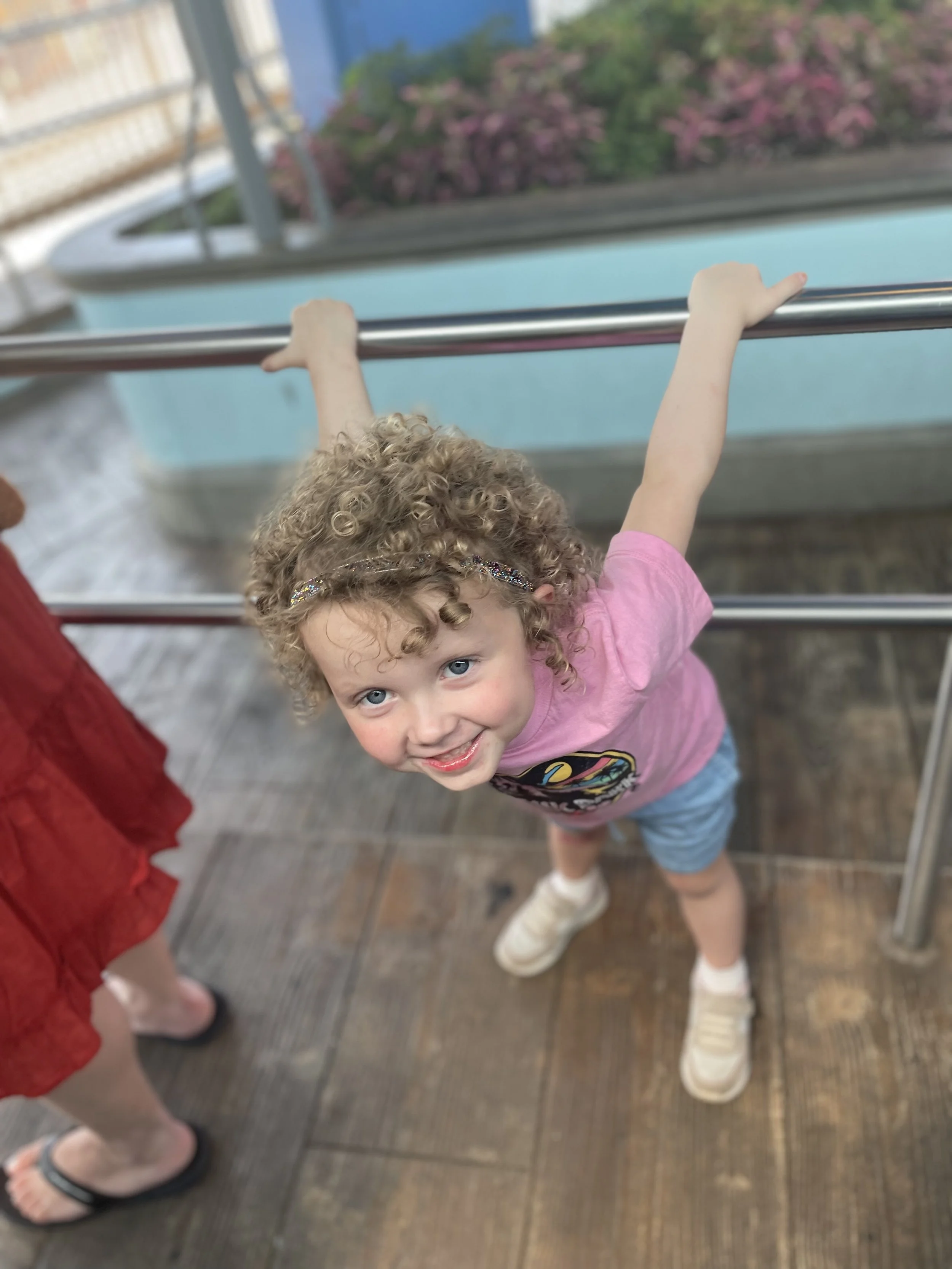 Young girl with curly hair and blue eyes smiling, wearing a pink t-shirt and shorts, holding onto a metal railing on a wooden deck.