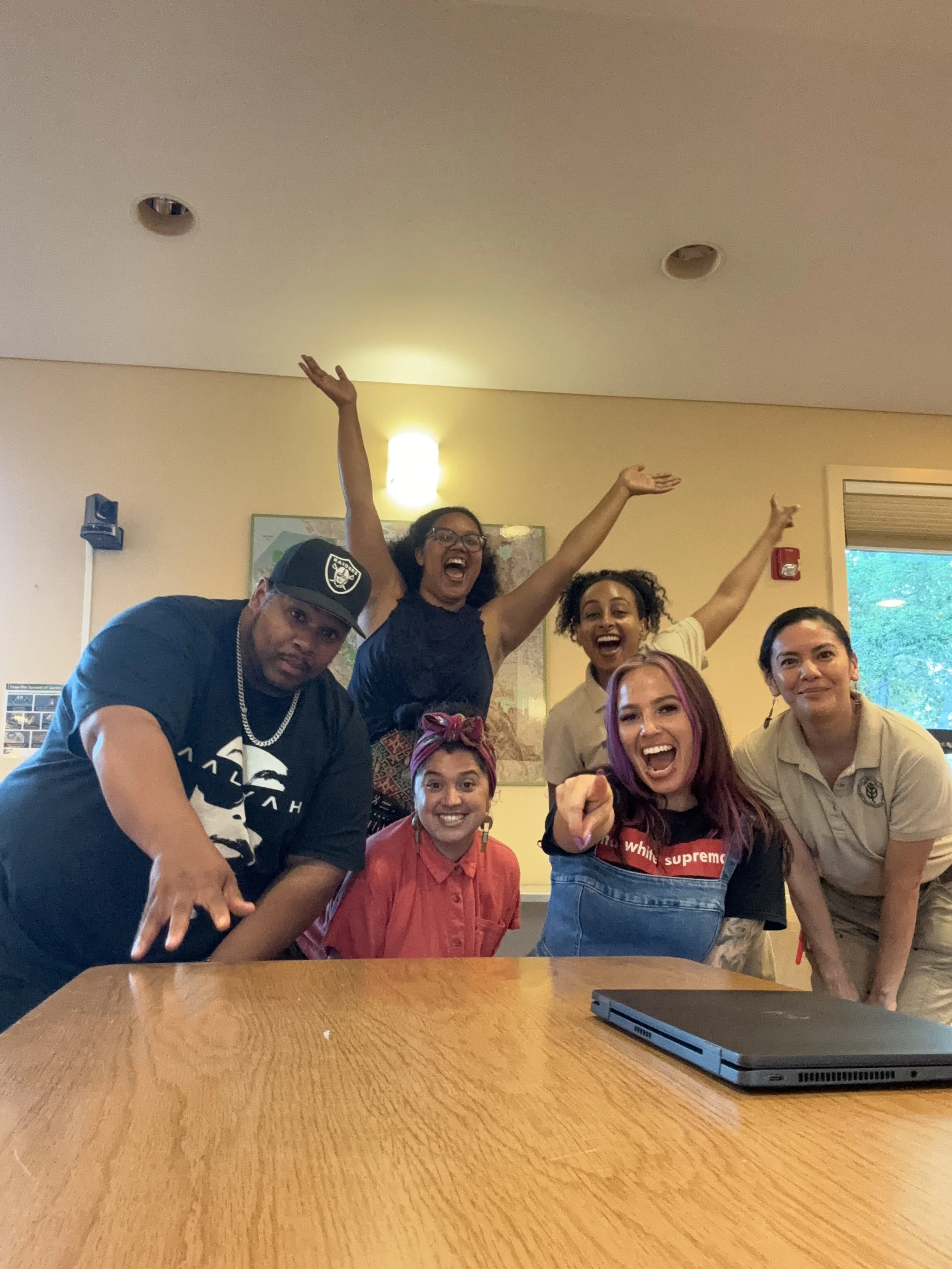 Six diverse people smiling and celebrating around a table in a room, with two women in the back raising their arms, one man leaning on the table pointing, and a woman in front pointing at the camera, with a laptop on the table.