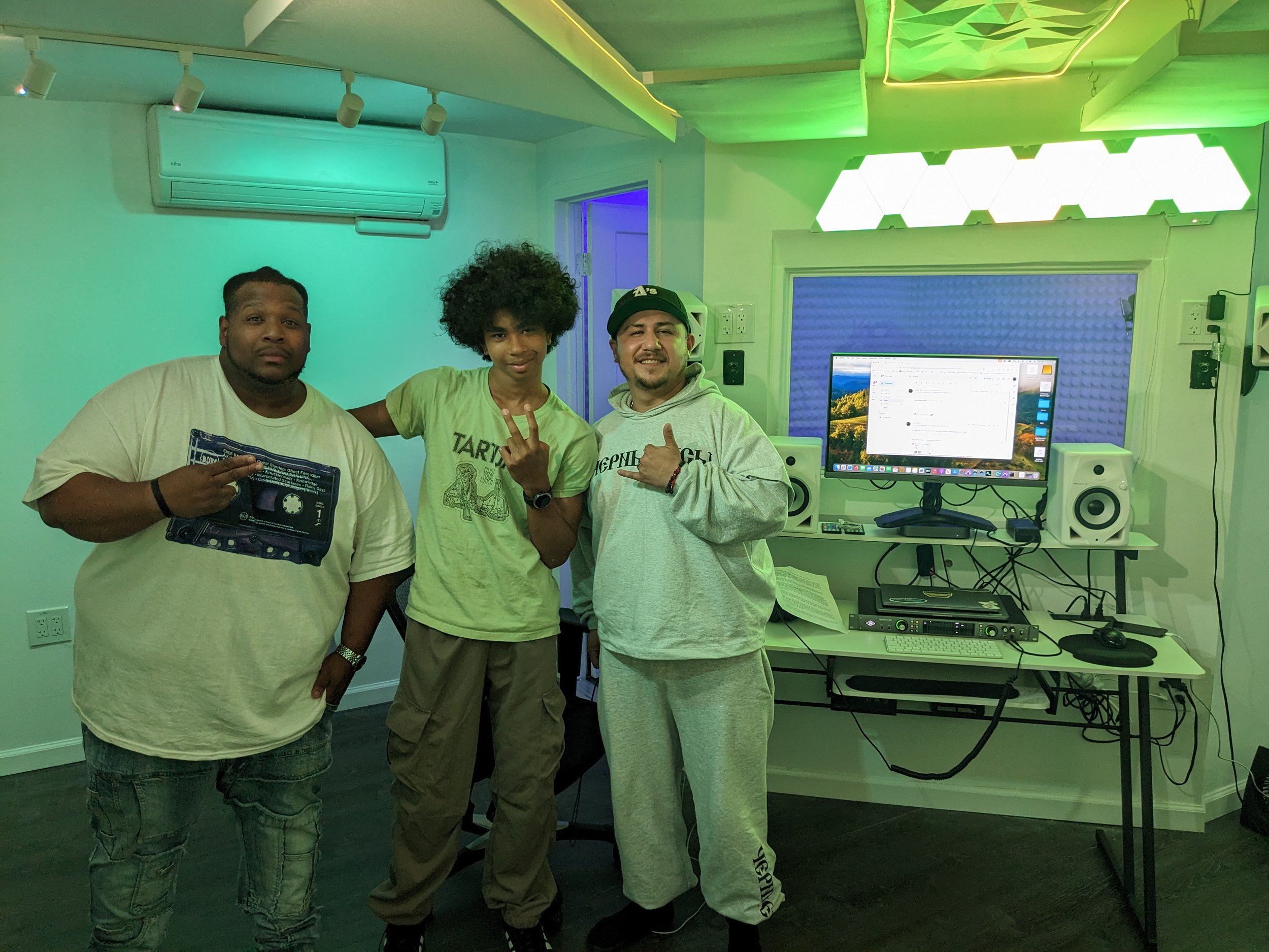 Three people standing in a music or recording studio, with one person holding a cassette tape, a computer monitor, speakers, and audio equipment behind them, with colorful lighting in the room.