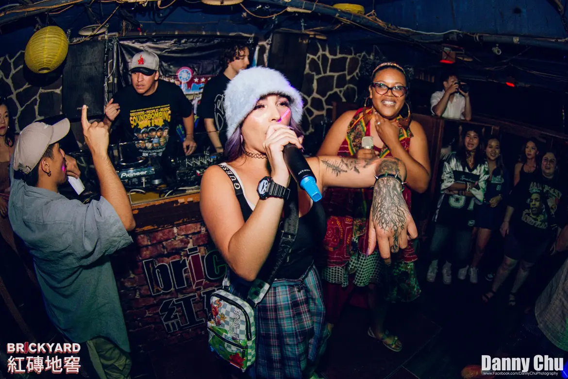 A woman singing into a microphone at a lively nightlife venue, with another woman beside her smiling, surrounded by people.