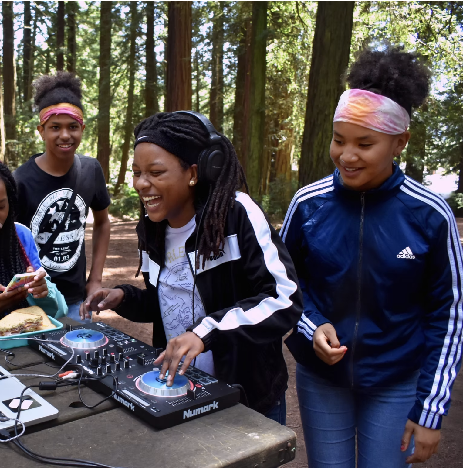 A group of women and girls in a forest, one woman is DJing with a DJ controller, others are watching and smiling, enjoying an outdoor event.