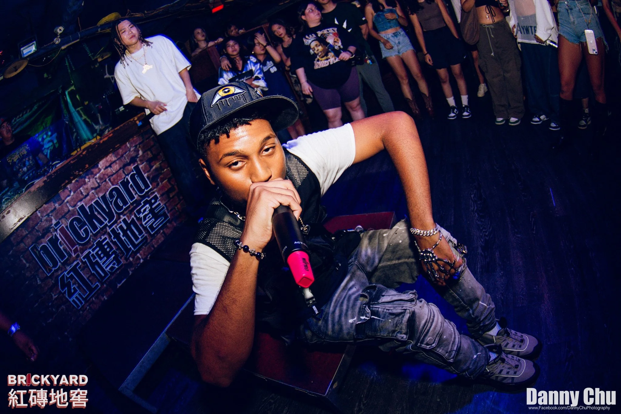 A young man holding a microphone and sitting on a brick platform at a crowded nightclub with a sign that reads 'BRICKYARD' in the background, surrounded by a group of people dancing and socializing.