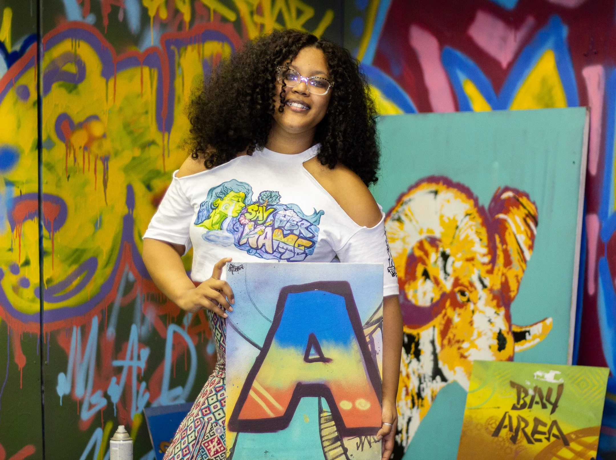 A woman with curly hair and glasses holding a colorful painting with the letter 'A' at an art event with graffiti art backgrounds and additional paintings.