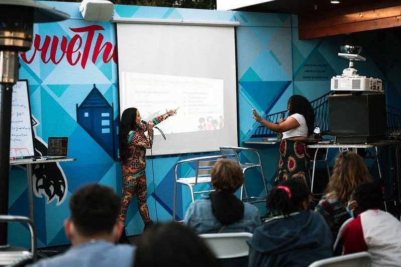 Two women are giving a presentation to an audience in a room with a blue geometric wall. One woman is speaking into a microphone, the other is pointing at the presentation screen. Audience members are seated, some wearing face masks.