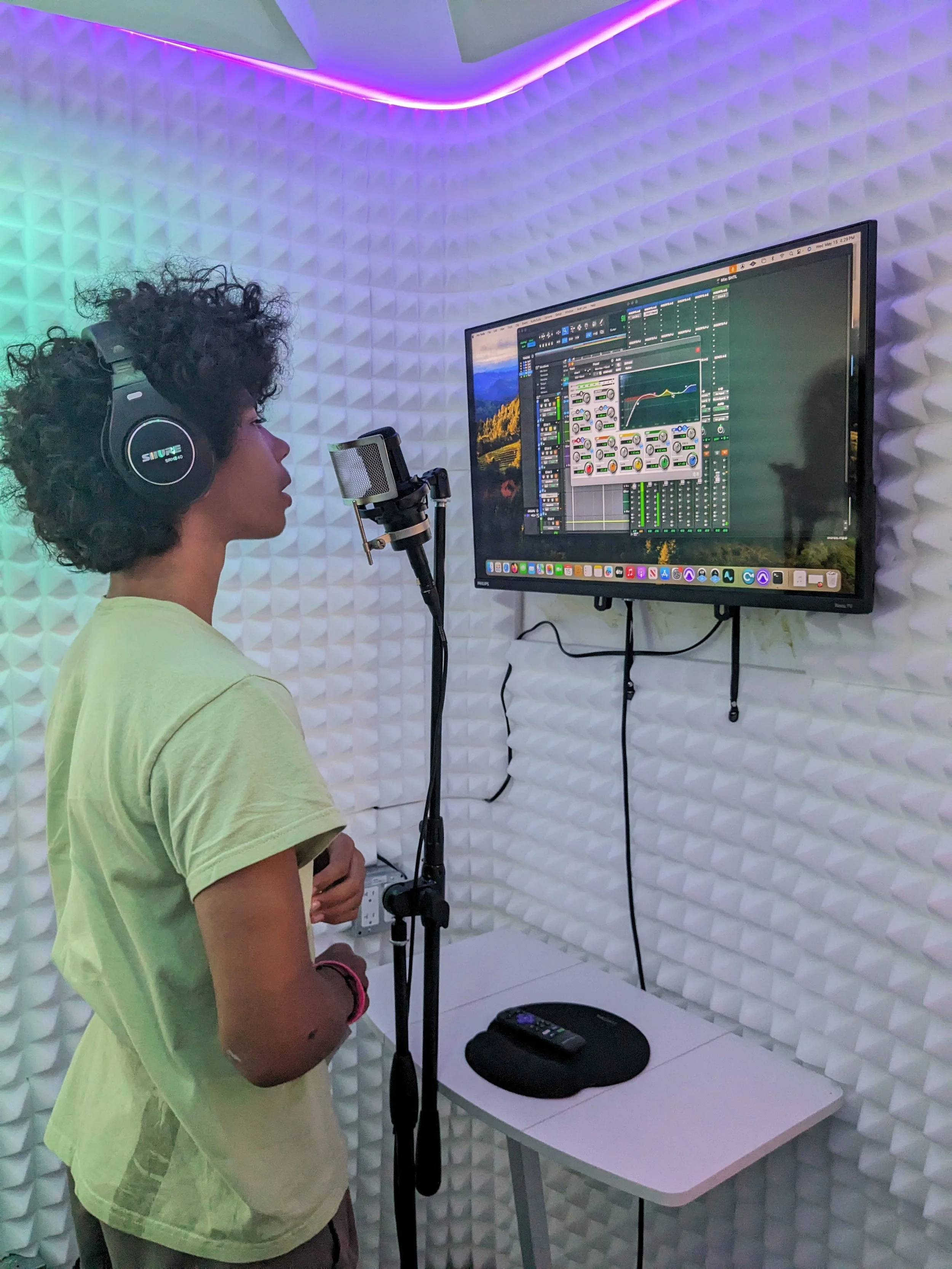 A person with curly hair wearing headphones stands in front of a microphone in a recording studio with padded white acoustic panels. They are facing a large screen displaying music production software, with a small table holding a remote control and a mouse.