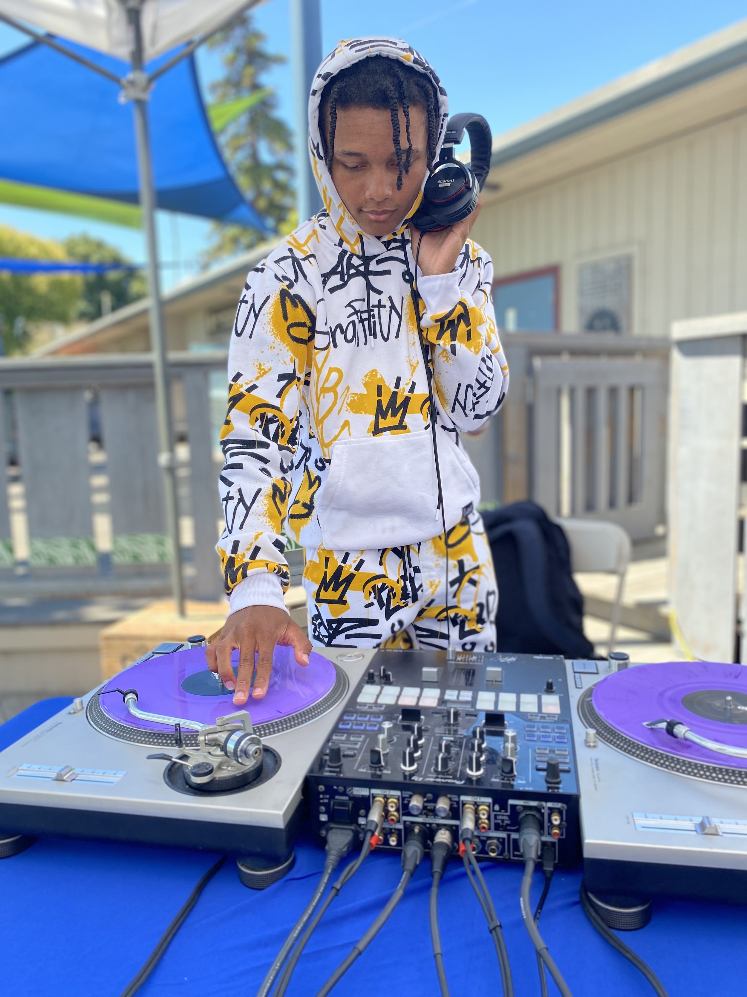 A young person with dreadlocks wearing a white hoodie and matching joggers with yellow and black graffiti patterns, standing outdoors on a deck, DJing with purple vinyl records on turntables, holding headphones in one ear, with a laptop and DJ controller on a blue table.