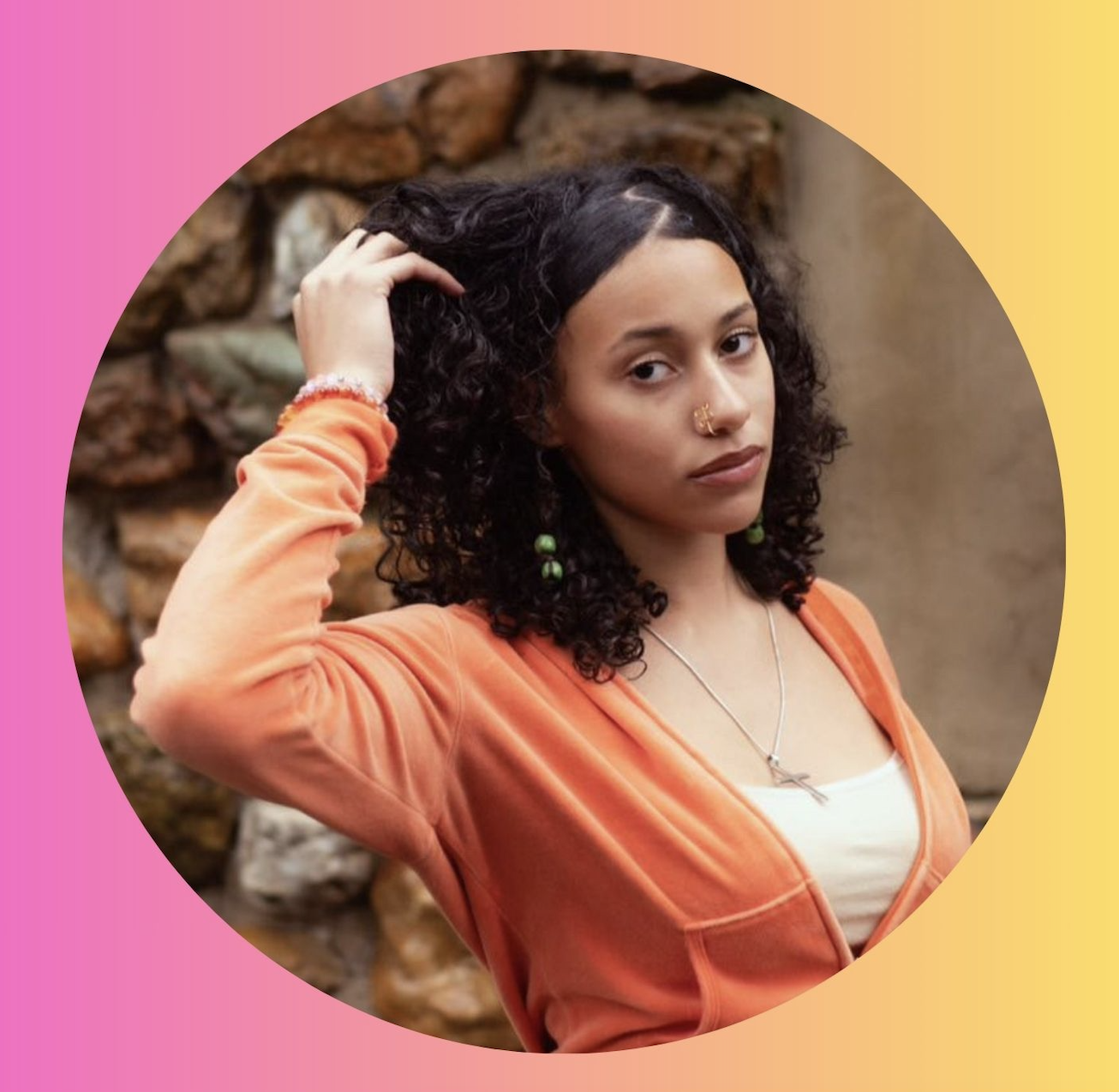 A young woman with curly dark hair, wearing an orange top and green earrings, gazes at the camera with a neutral expression, standing outdoors against a stone wall background.
