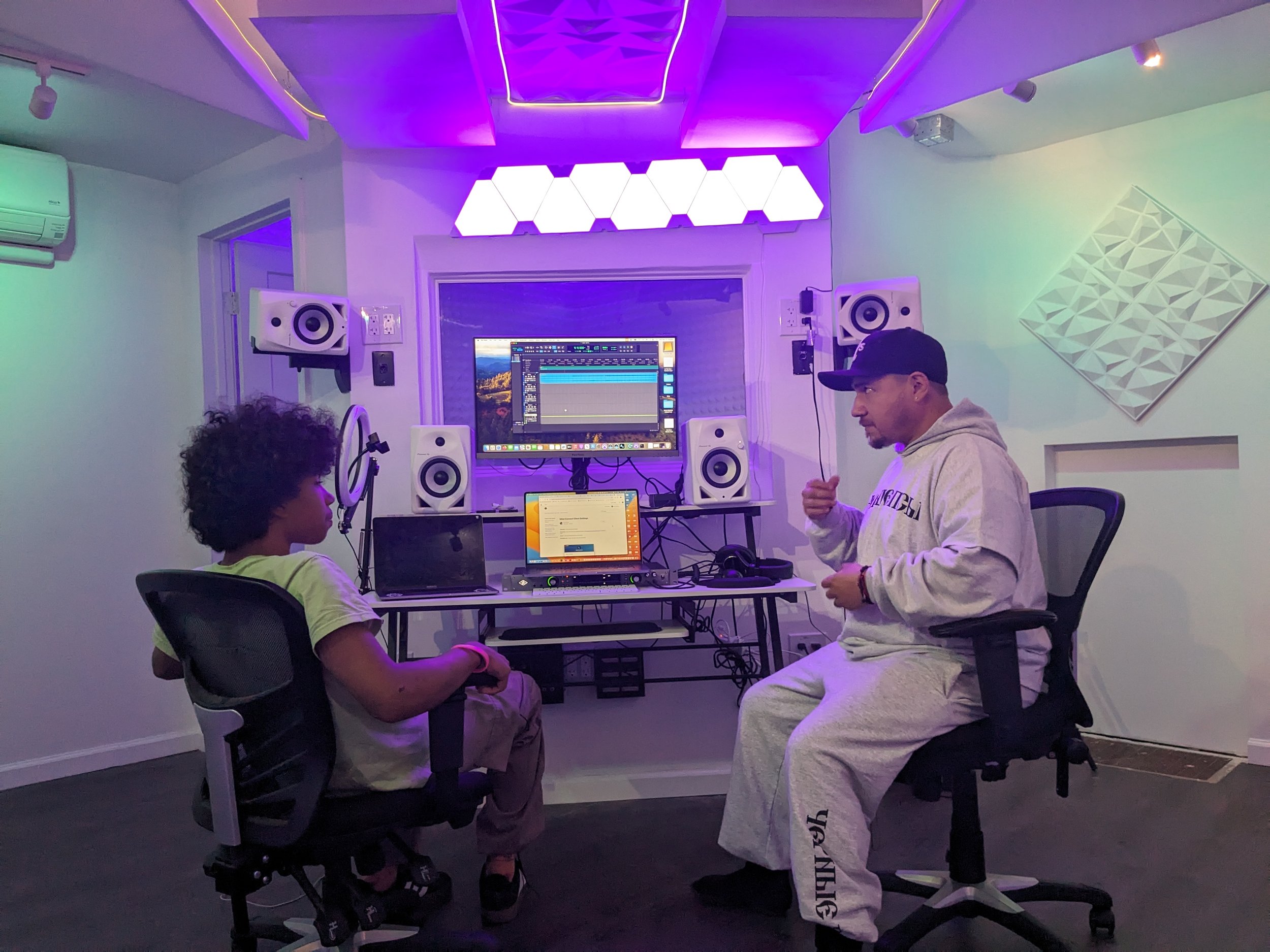 A young boy and an adult man sitting in a music studio with studio monitors, a computer, and sound equipment, illuminated by colorful LED lights.