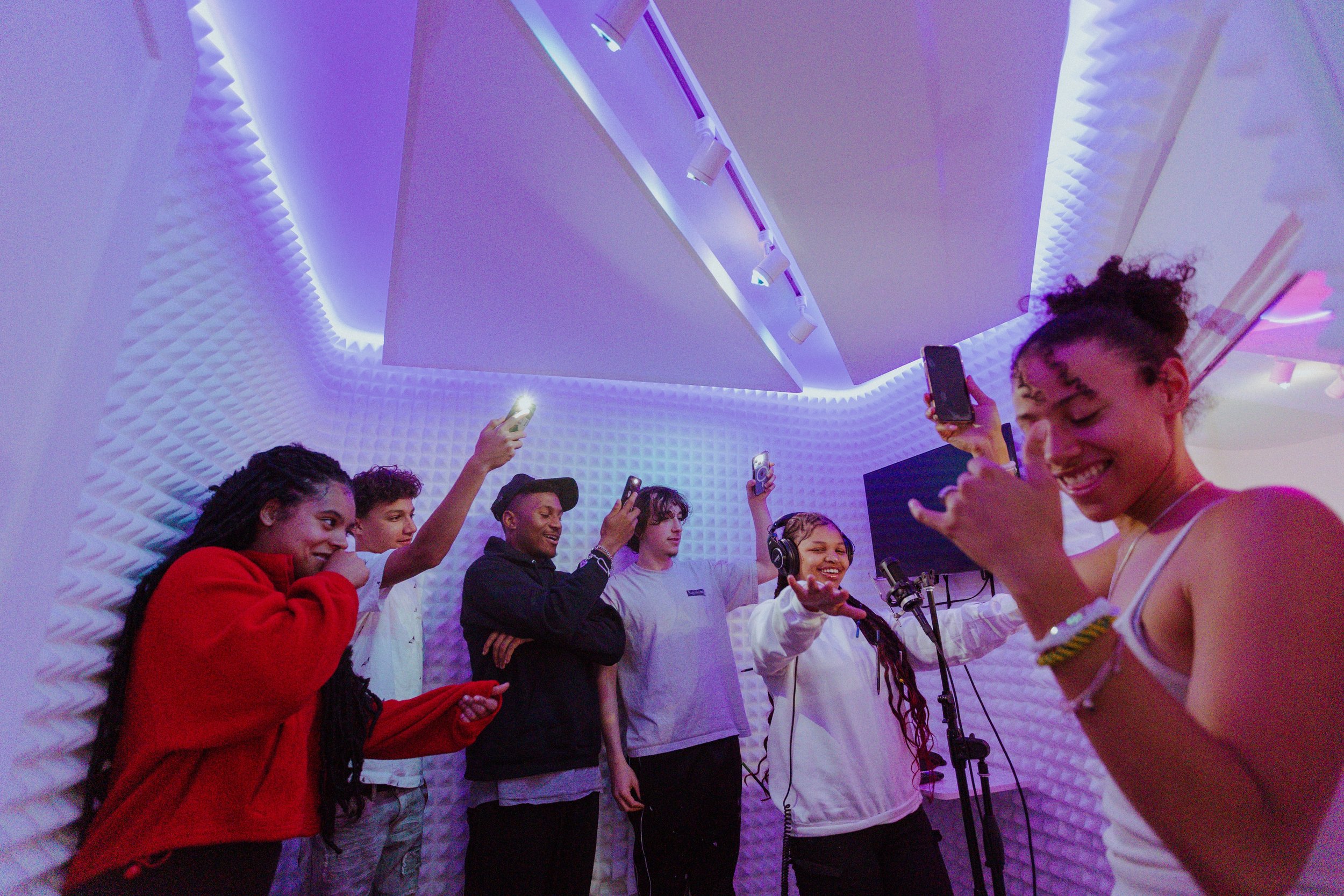 A group of six young people in a recording studio, some recording vocals and others taking photos or videos with their phones, surrounded by white soundproofing panels with purple lighting.