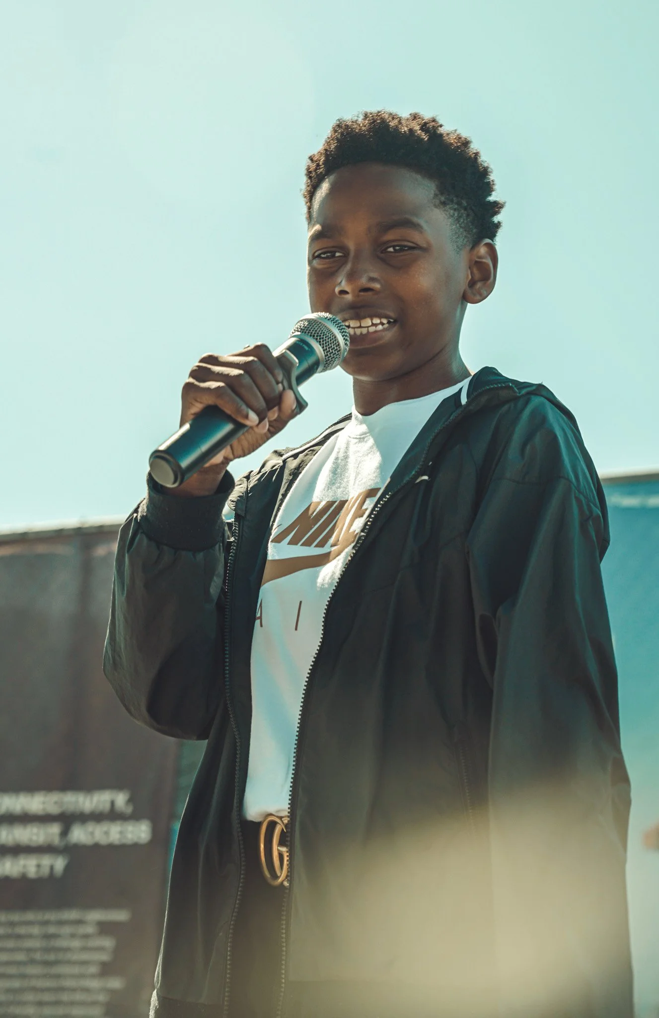 A young man speaking into a microphone outdoors, wearing a black jacket over a white Nike t-shirt, with a blurred outdoor background.
