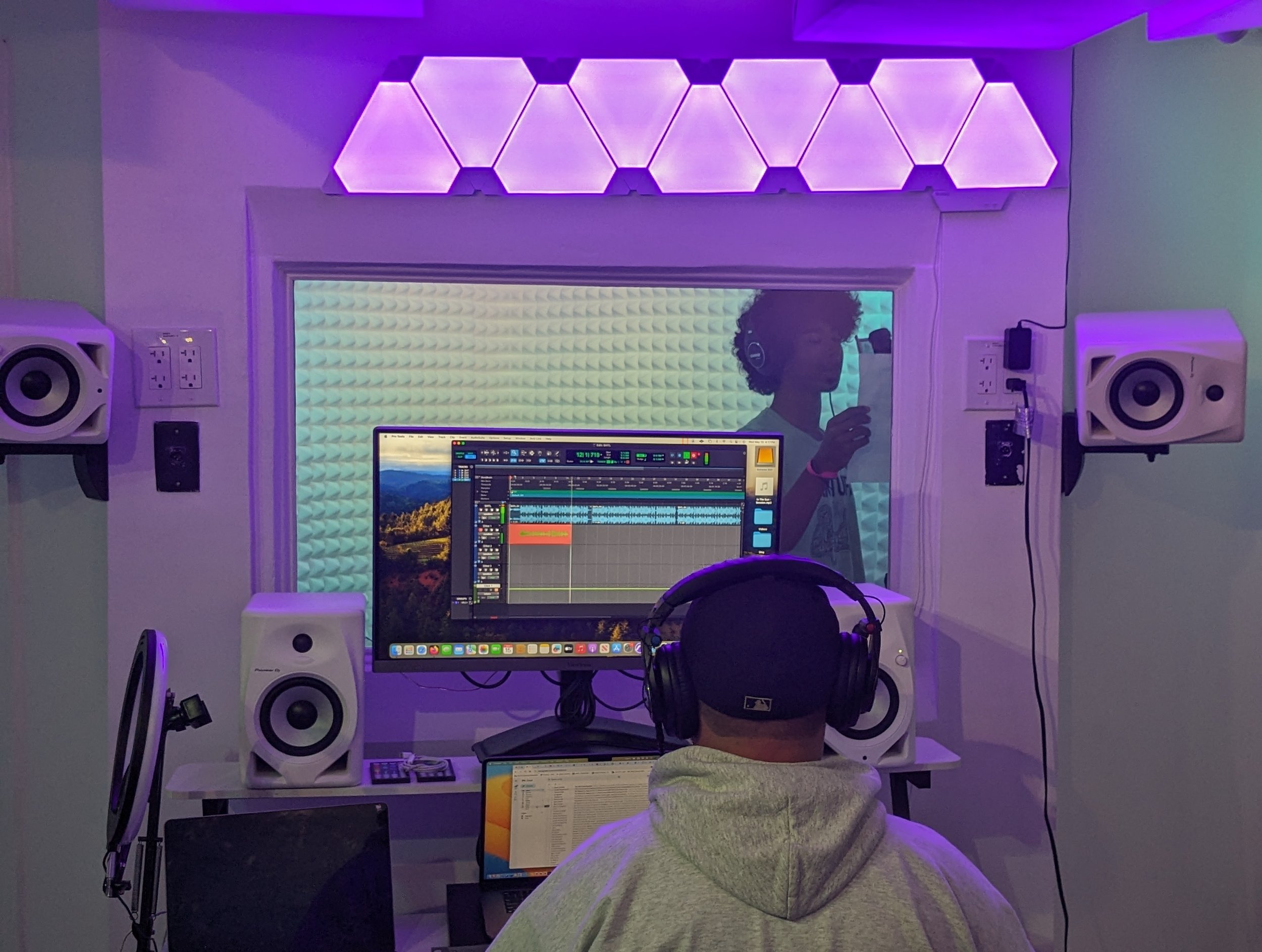 A person in a hoodie and baseball cap wearing headphones sitting in front of a computer with music production software, in a studio with purple lighting and soundproofing panels.