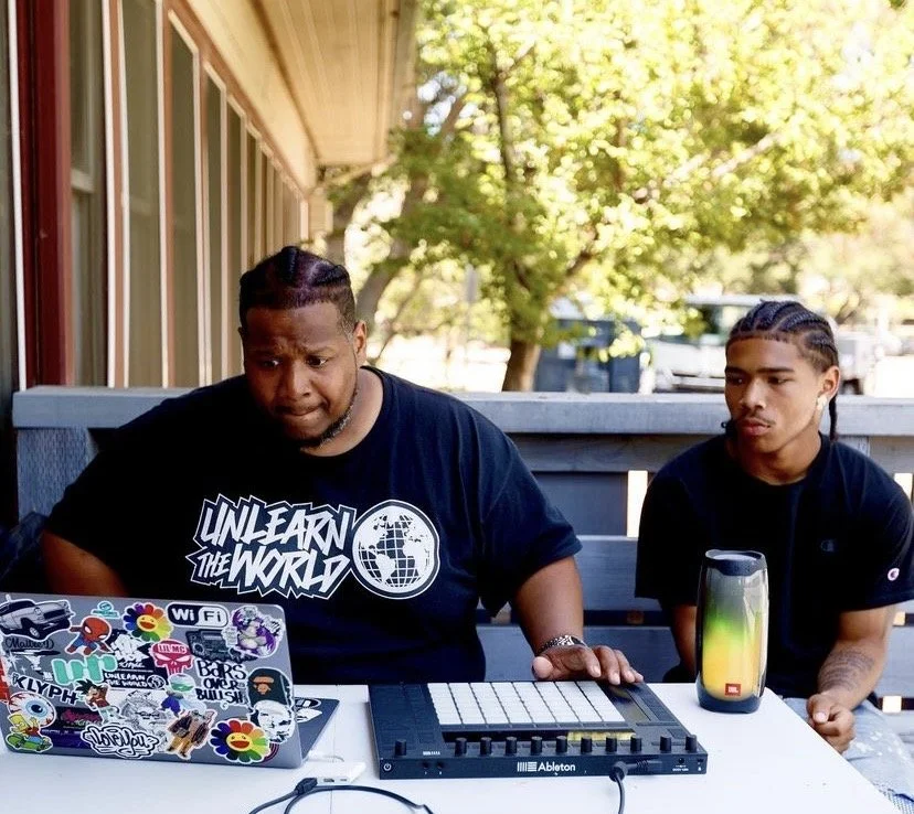 Two young men sitting at a table outdoors with music equipment, one working on a laptop covered in stickers and the other observing, during daytime with trees in the background.