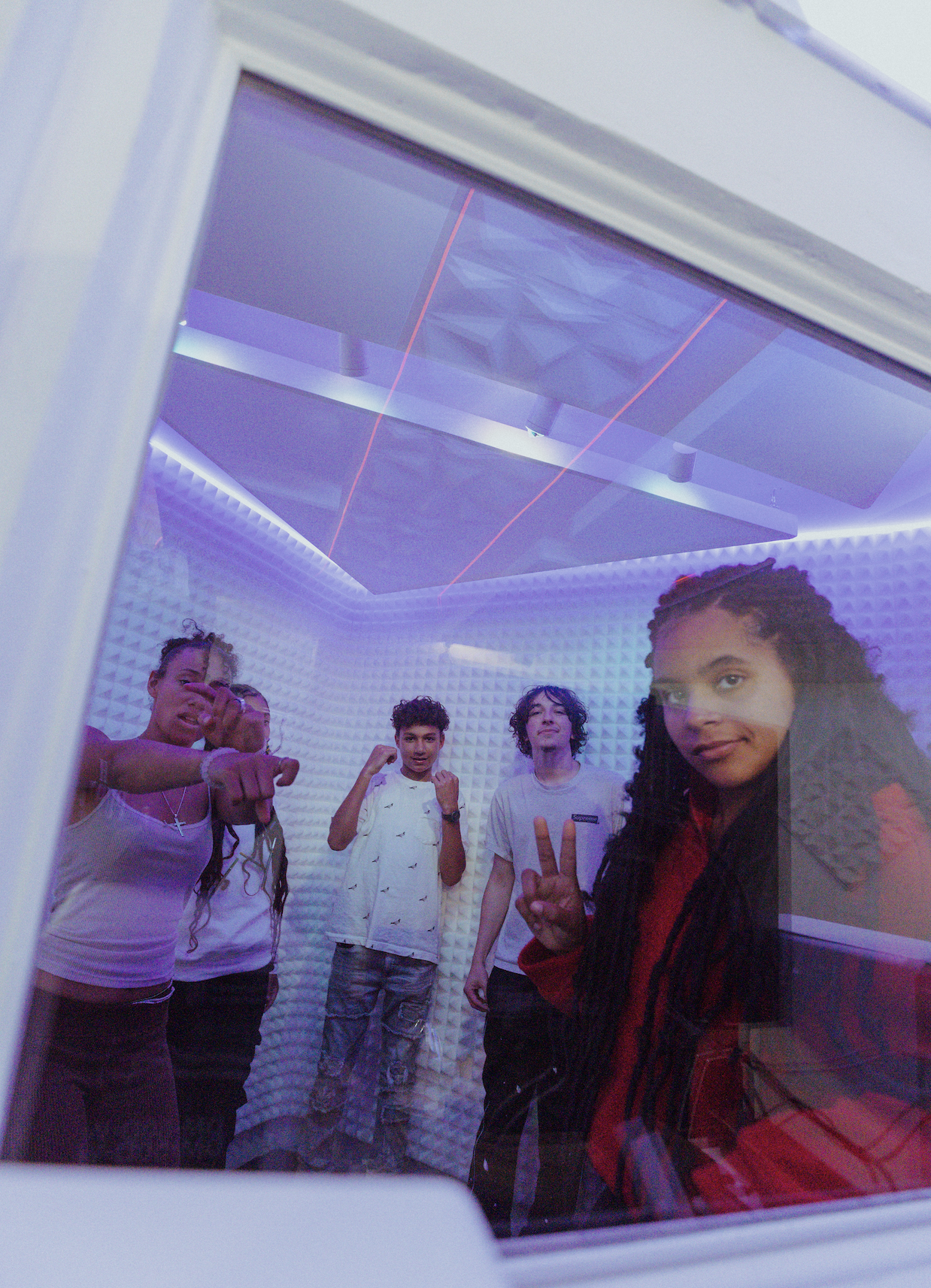 A group of five young people in a recording studio, seen through a glass window, with one making a peace sign and others posing playfully.