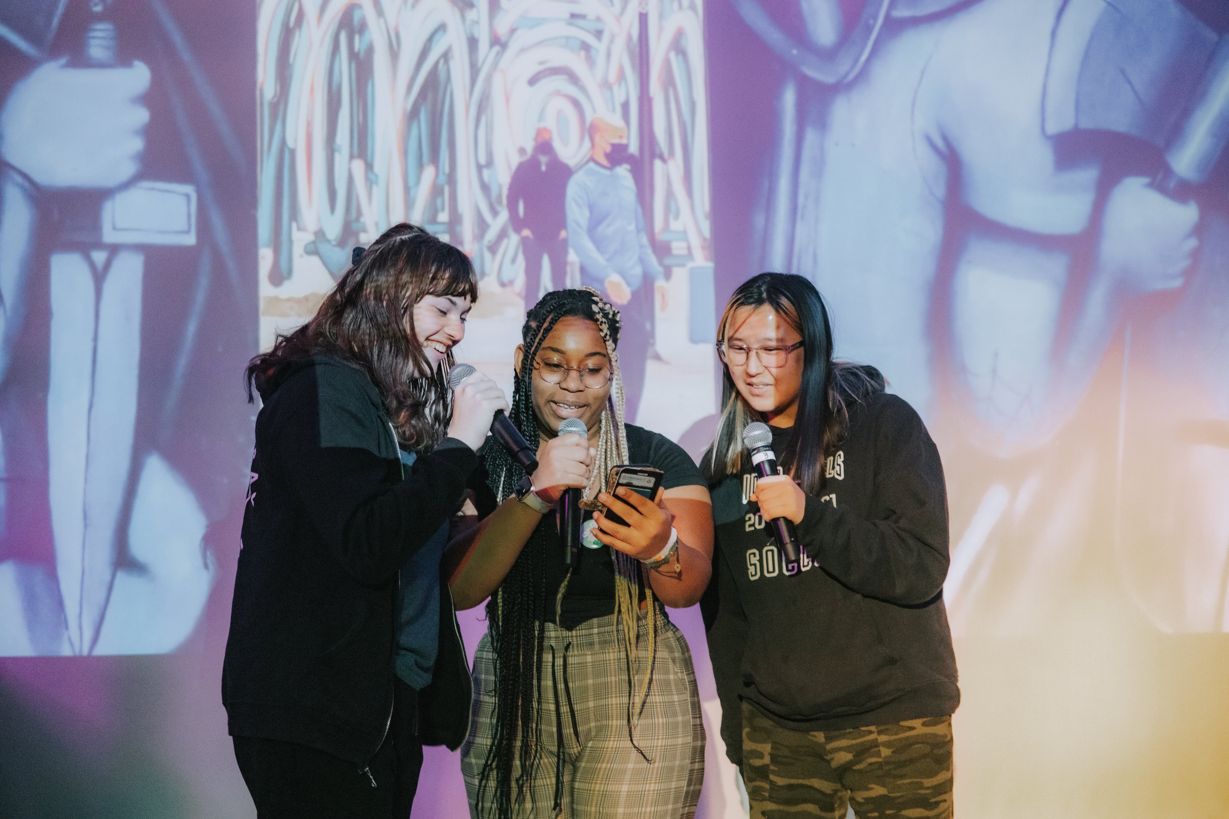 Three women standing closely together, holding microphones and looking at a smartphone, on stage with a colorful abstract mural in the background.