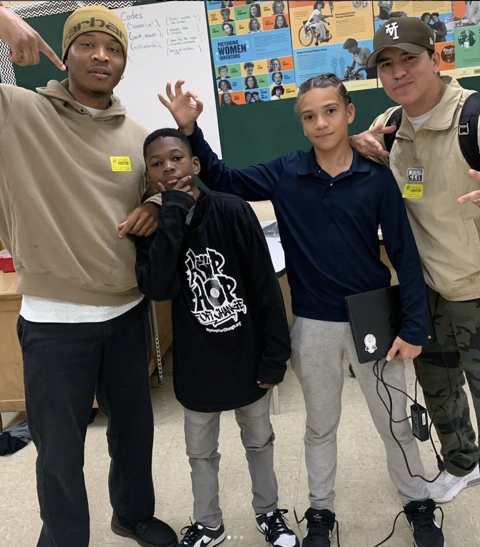 Two adult men and two boys in a classroom. The men are wearing beige hoodies with visitor tags, and the boys are in casual clothing. The background shows educational posters and a whiteboard with notes.