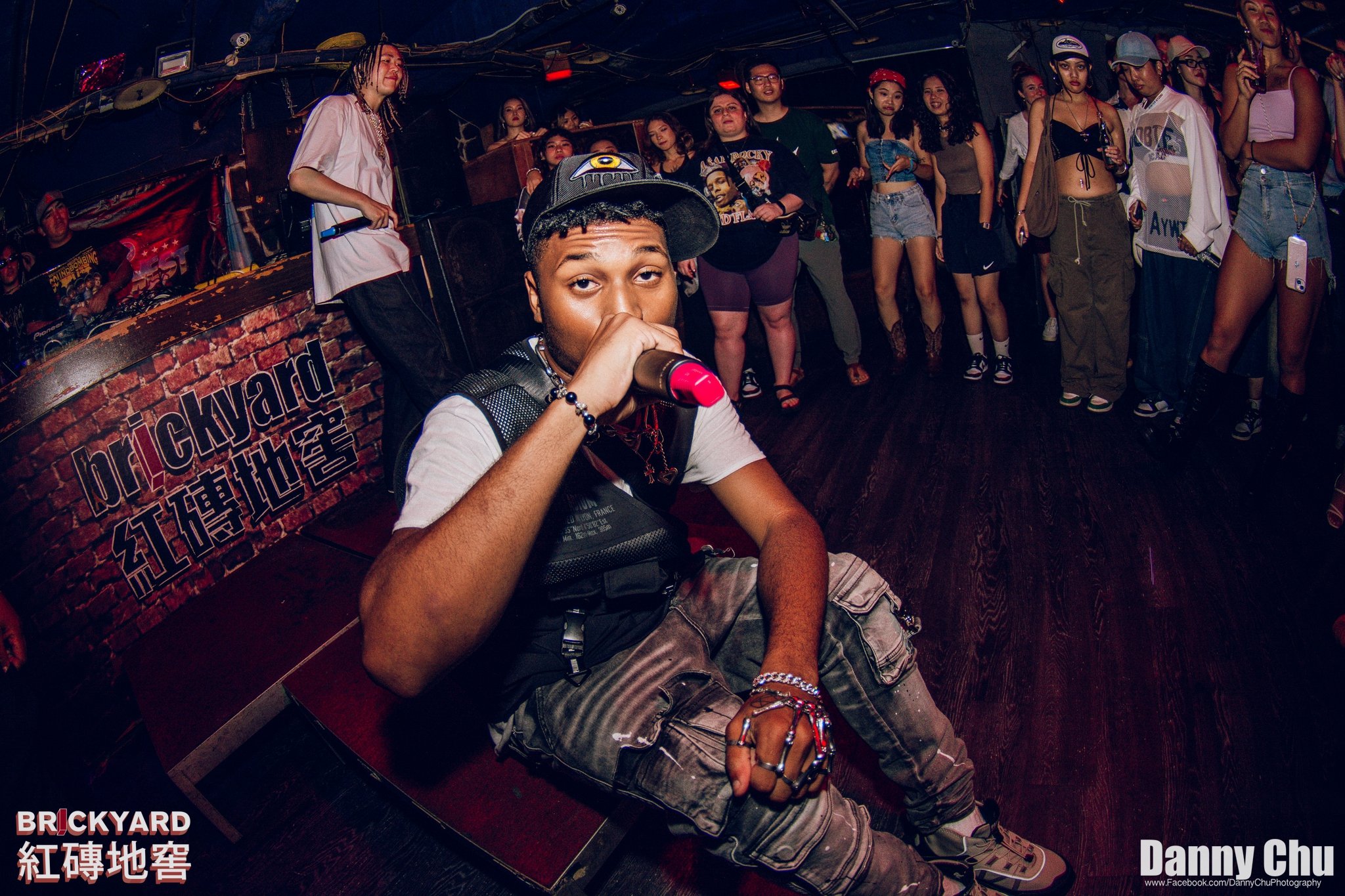 A young man holding a microphone on stage with a group of women in the background at a music venue called Brickyard.