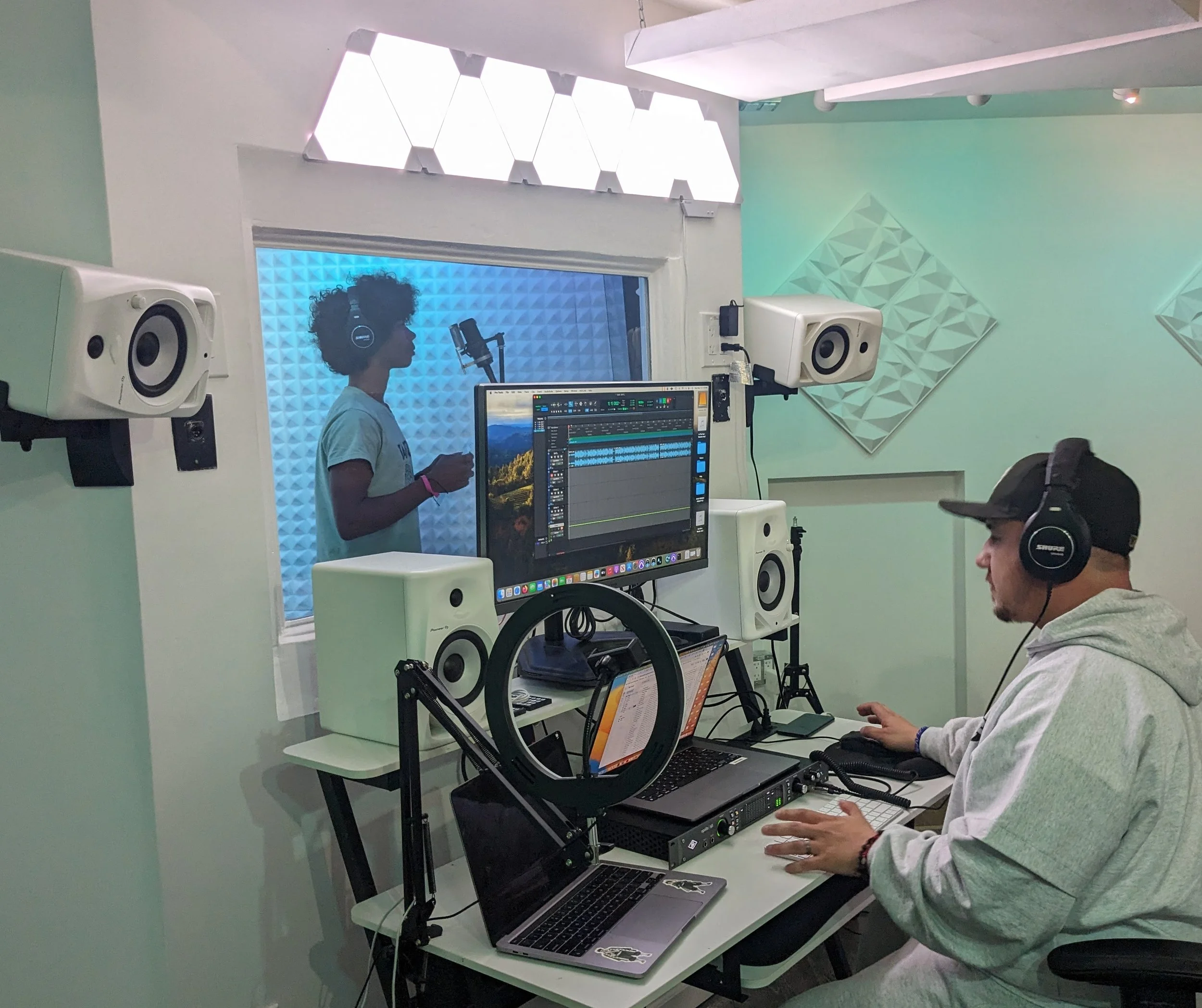 A music recording studio with two people: a woman in the recording booth singing into a microphone, and a man outside the booth operating audio equipment. Both are wearing headphones.