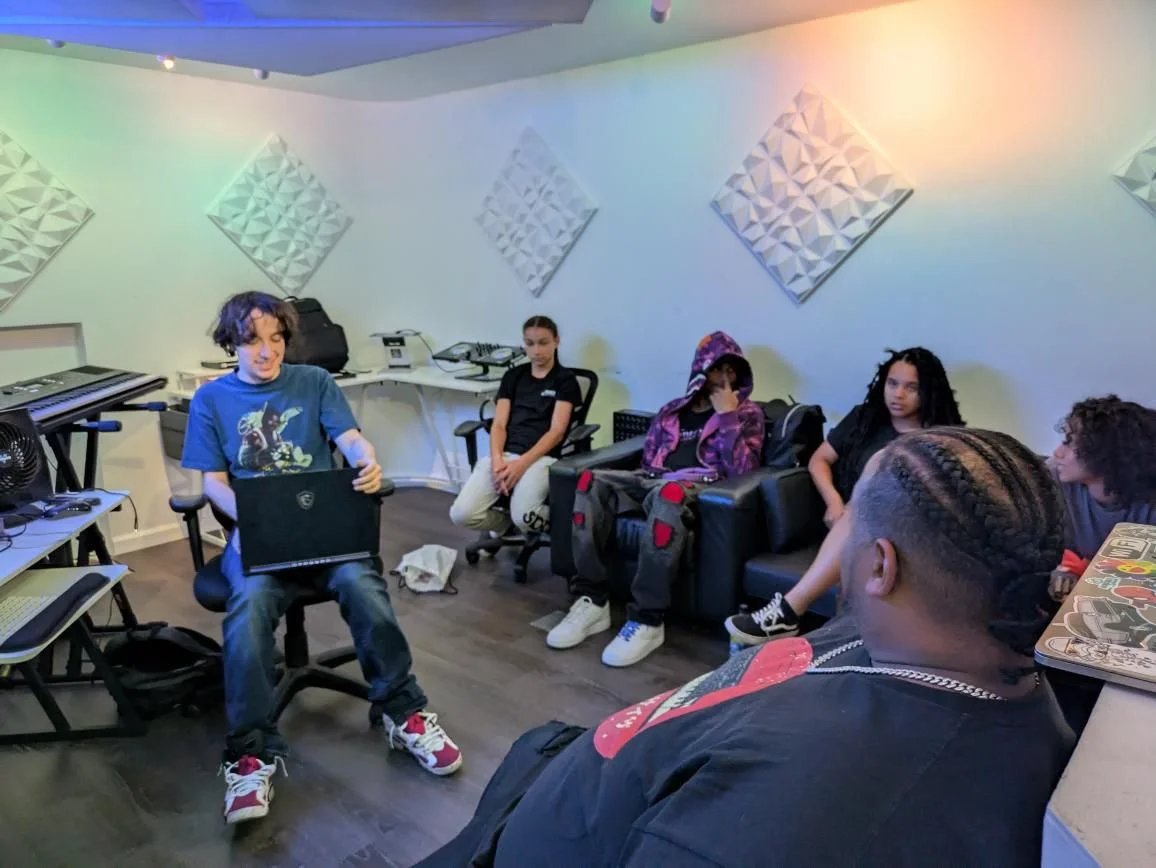 Group of five young people in a music studio or classroom, sitting on chairs and a sofa, with electronic music equipment and acoustic panels on the wall.