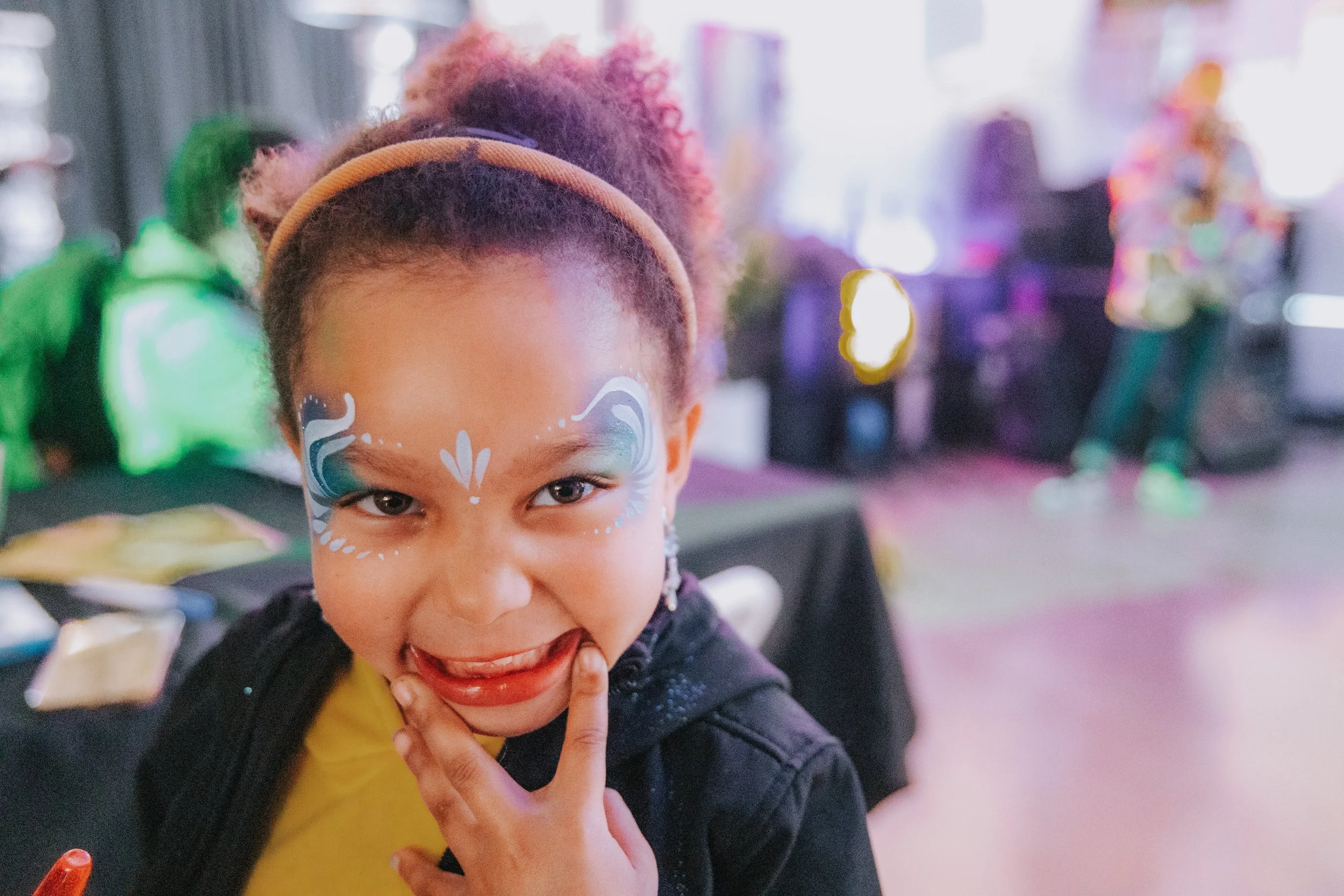 A young girl with face paint and a yellow shirt, smiling playfully with her finger near her mouth, in a colorful indoor setting.