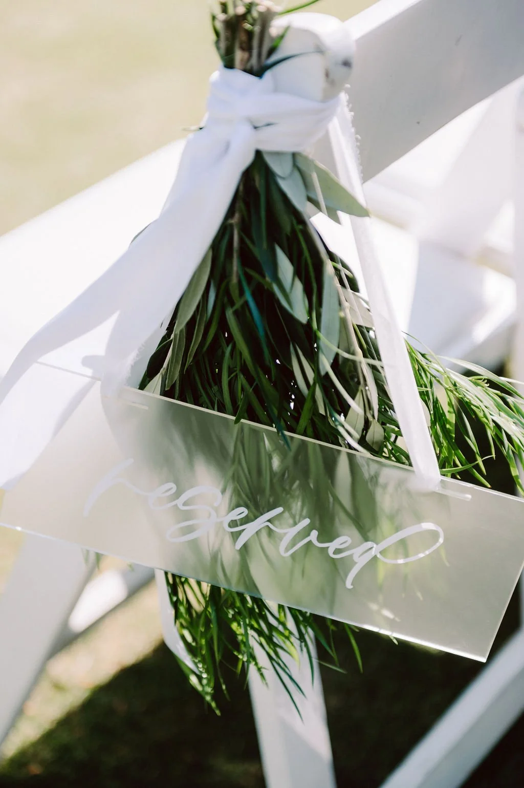 Bouquet of green leaves tied with white ribbon on a white chair, with a transparent sign reading 'Reserved' attached.