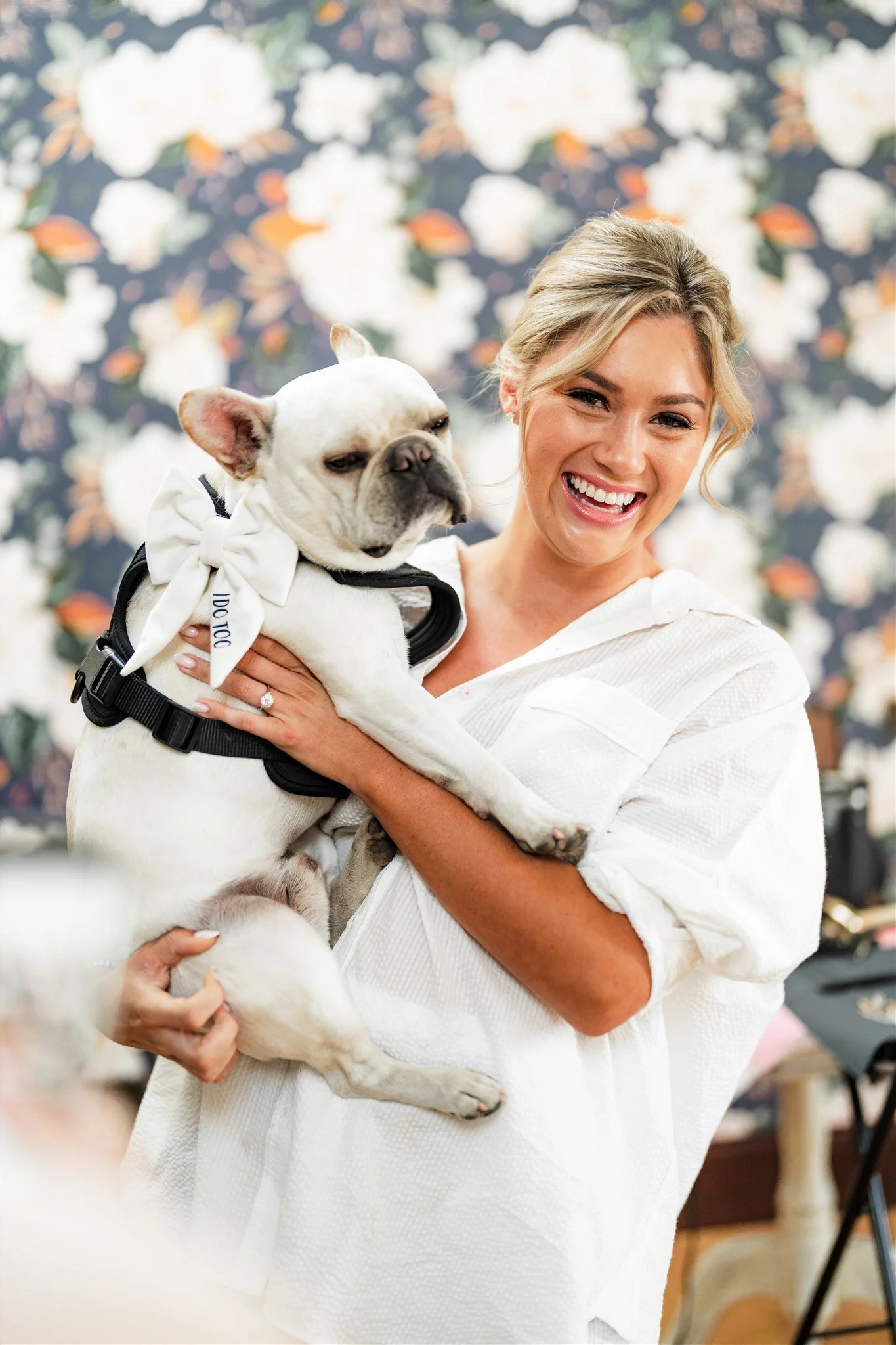 A woman with blonde hair smiling and holding a French bulldog puppy with a white bow around its neck, against a floral patterned background.