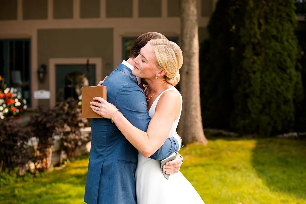 A bride and groom embrace and share a hug outside a house, with the bride holding a small book.