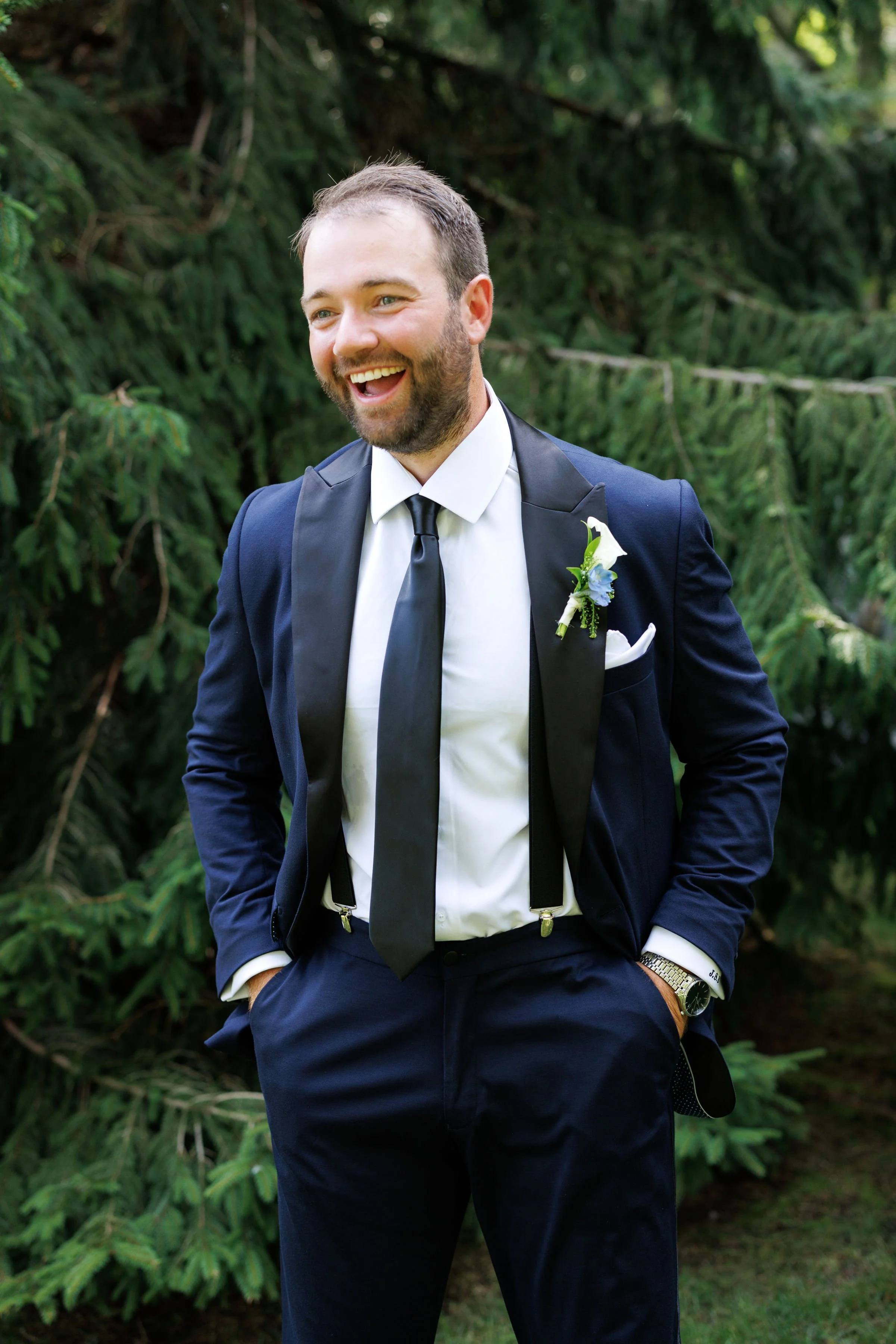 A smiling man in a tuxedo with suspenders and a boutonnière, standing outdoors in front of green trees.