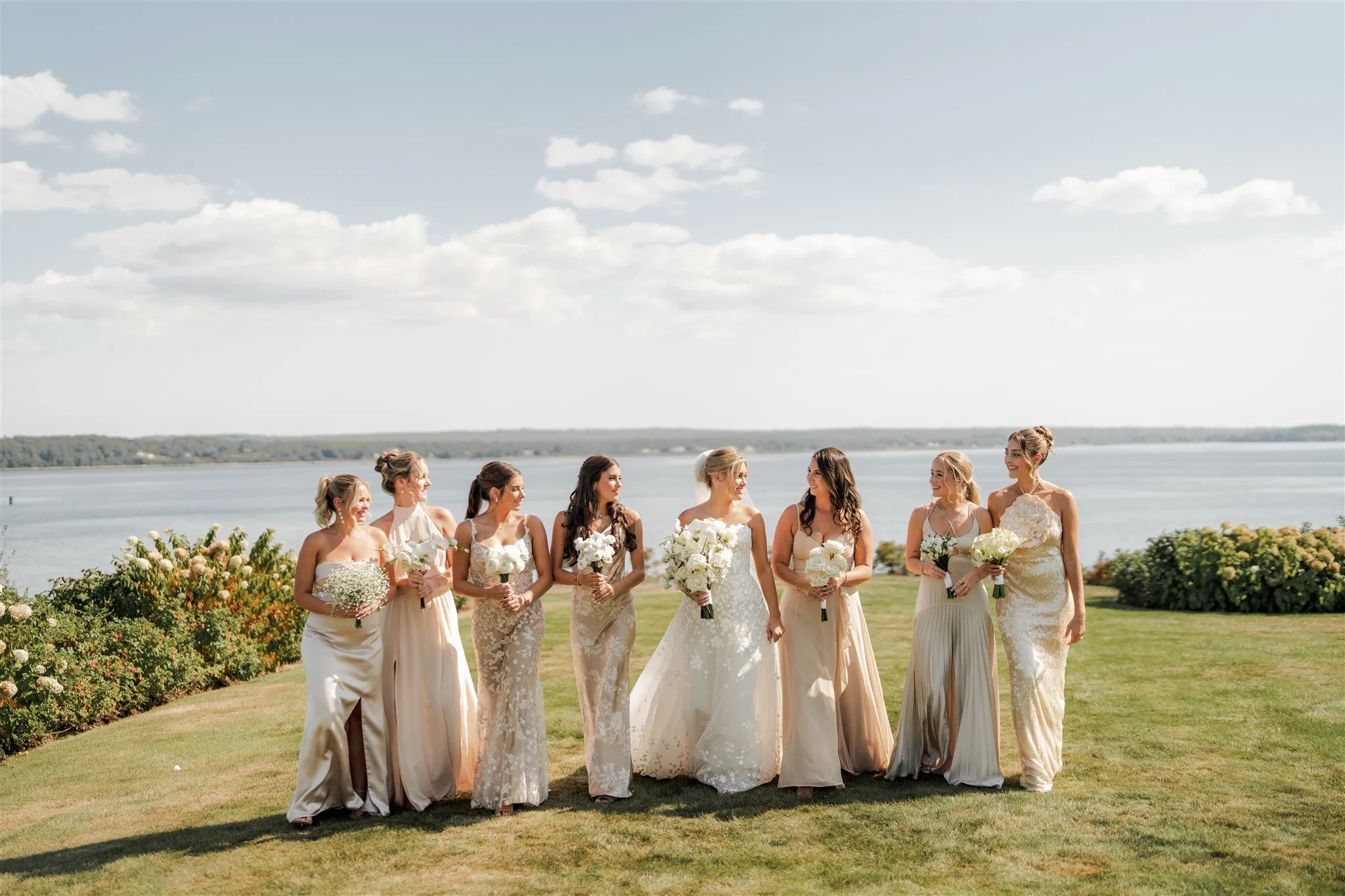 Group of women in formal dresses standing outdoors near a body of water, holding bouquets of flowers, with a cloudy sky in the background.