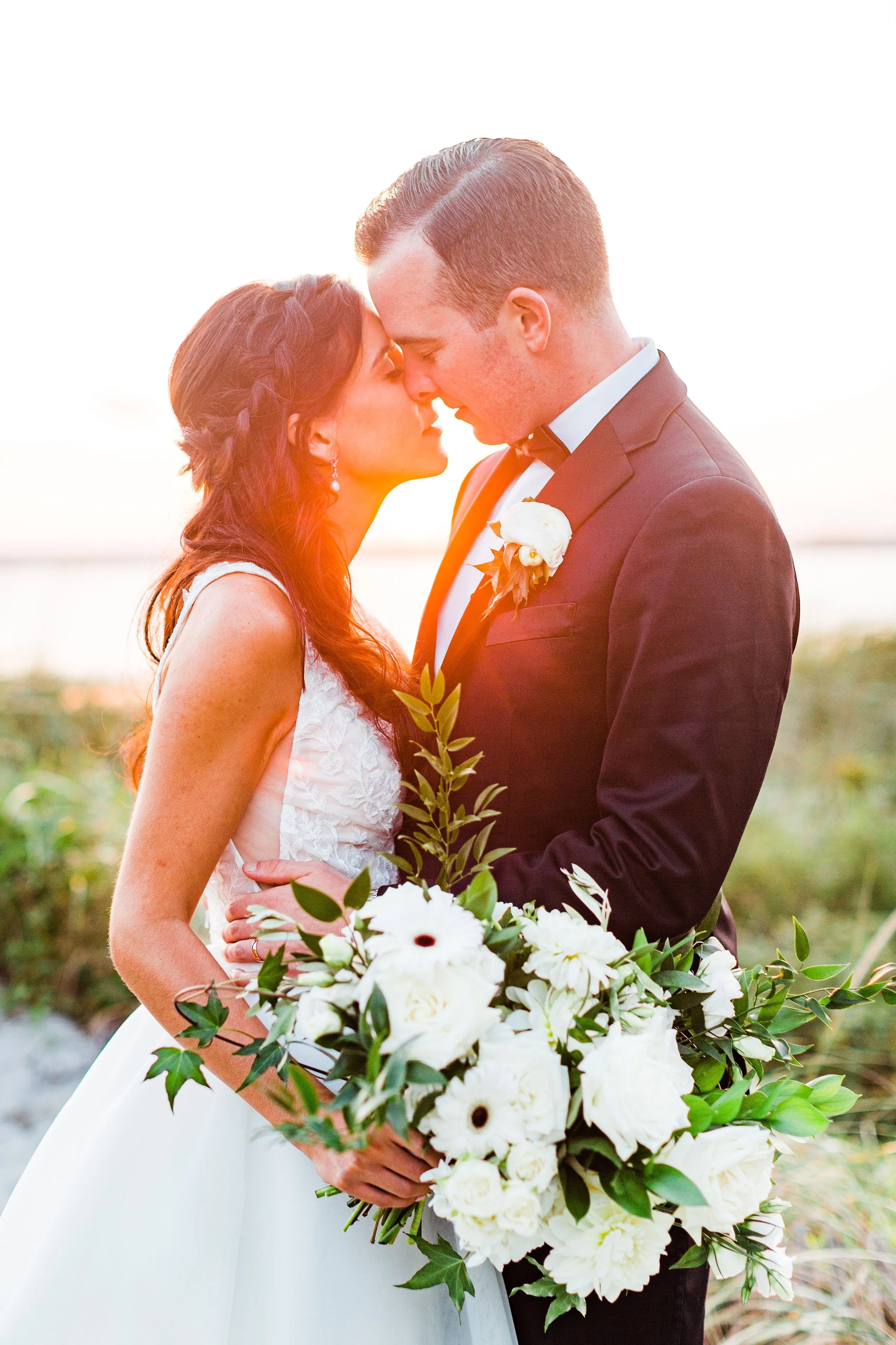 A wedding couple, dressed in formal attire, stands close with foreheads touching, outdoors at sunset, holding a large bouquet of white flowers and greenery.
