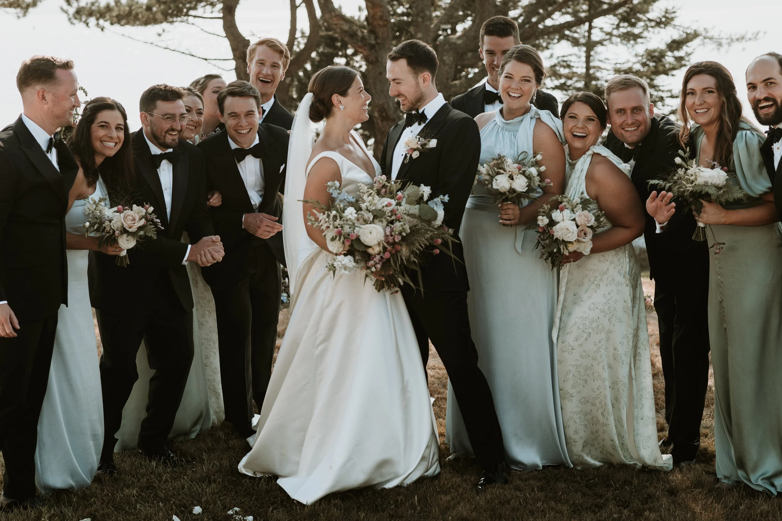A wedding party outdoors with a bride and groom in the center, surrounded by friends and family smiling and holding bouquets, in a natural setting with trees in the background.