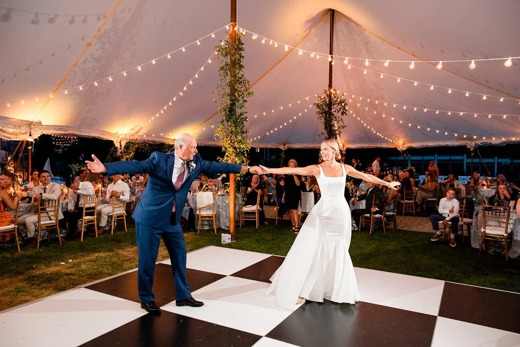 A bride and an older man dancing together at a wedding reception under a decorated tent with string lights, surrounded by seated guests.