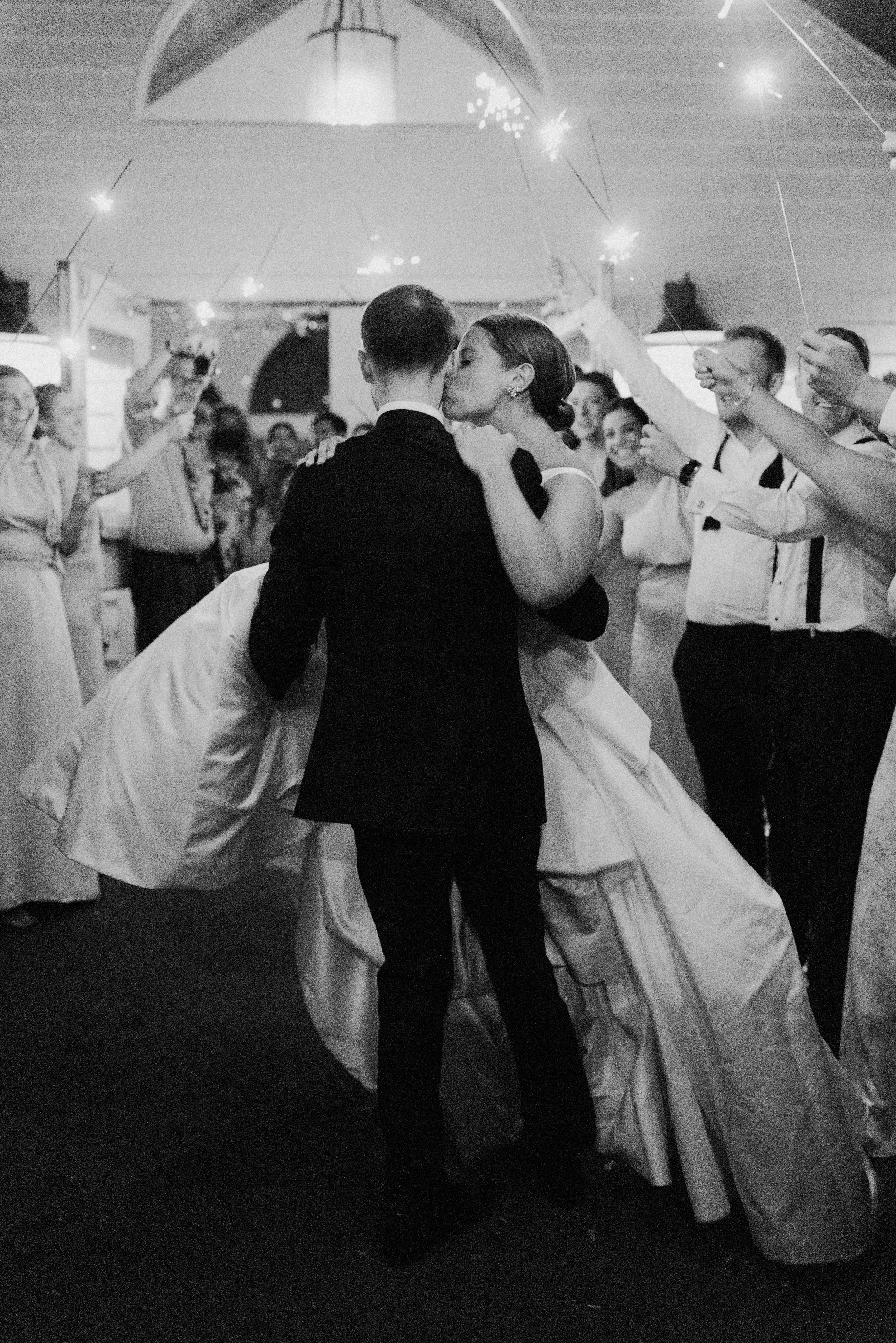 A bride and groom share a kiss on the dance floor during their wedding reception, surrounded by guests holding sparklers.
