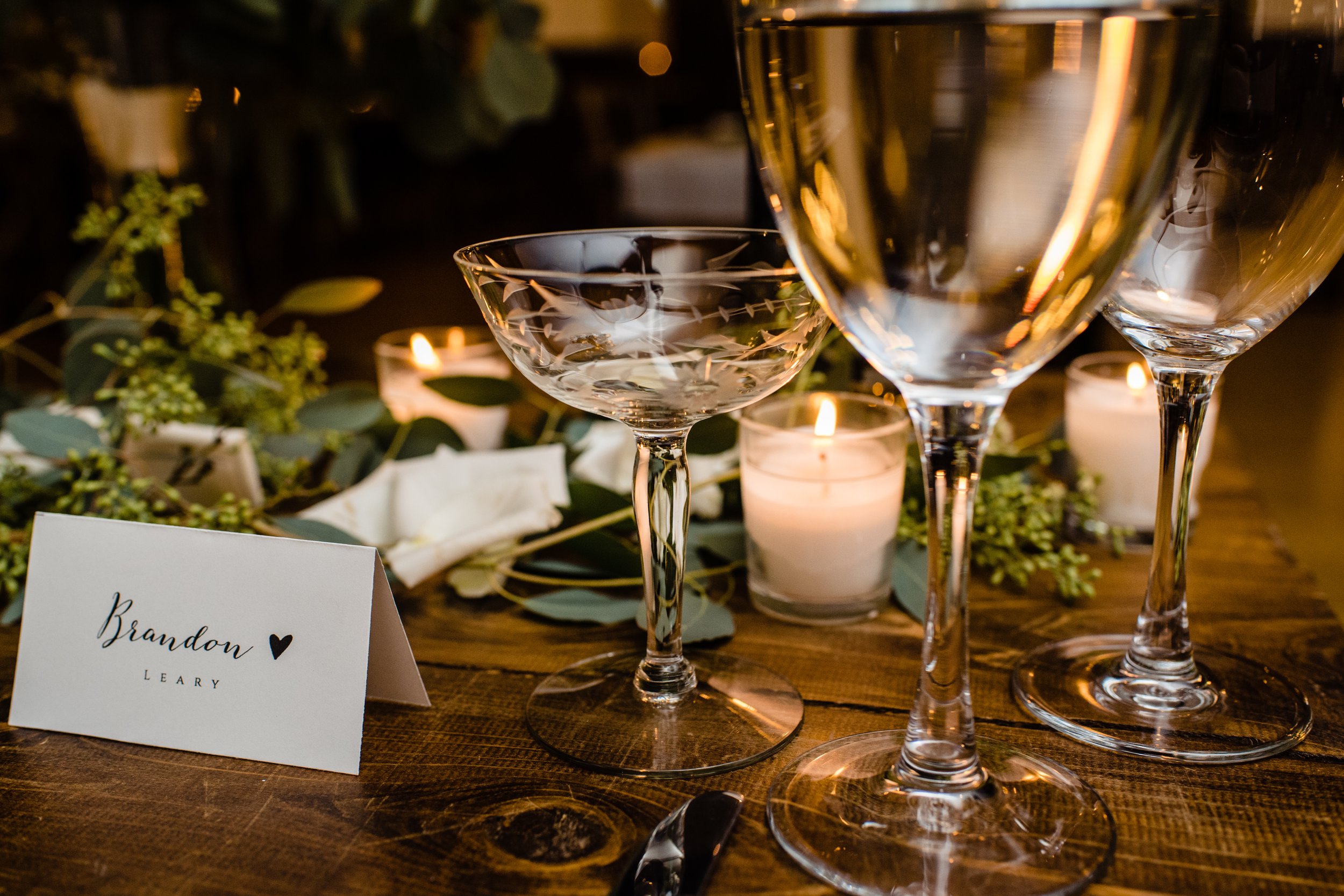 Elegant wedding reception table with three champagne glasses, candles, greenery, and a place card for Brandon Leary.