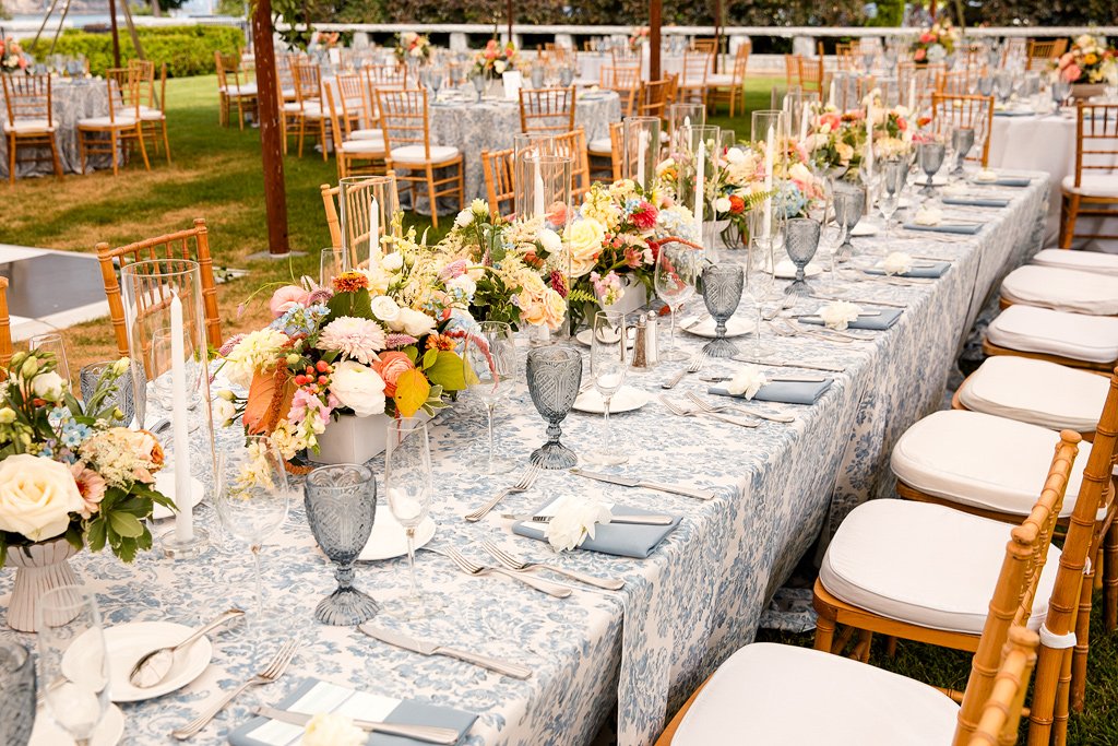 Long banquet table with floral centerpieces, glassware, and table settings at an outdoor event reception.