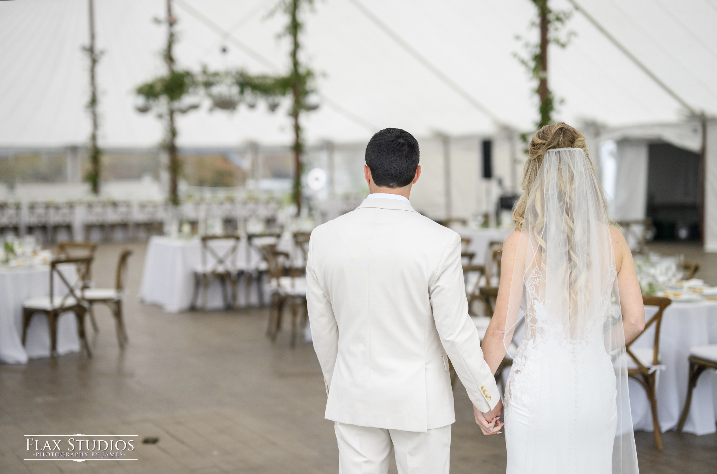 A bride and groom hold hands while walking hand in hand inside a decorated wedding venue with tables and chairs.