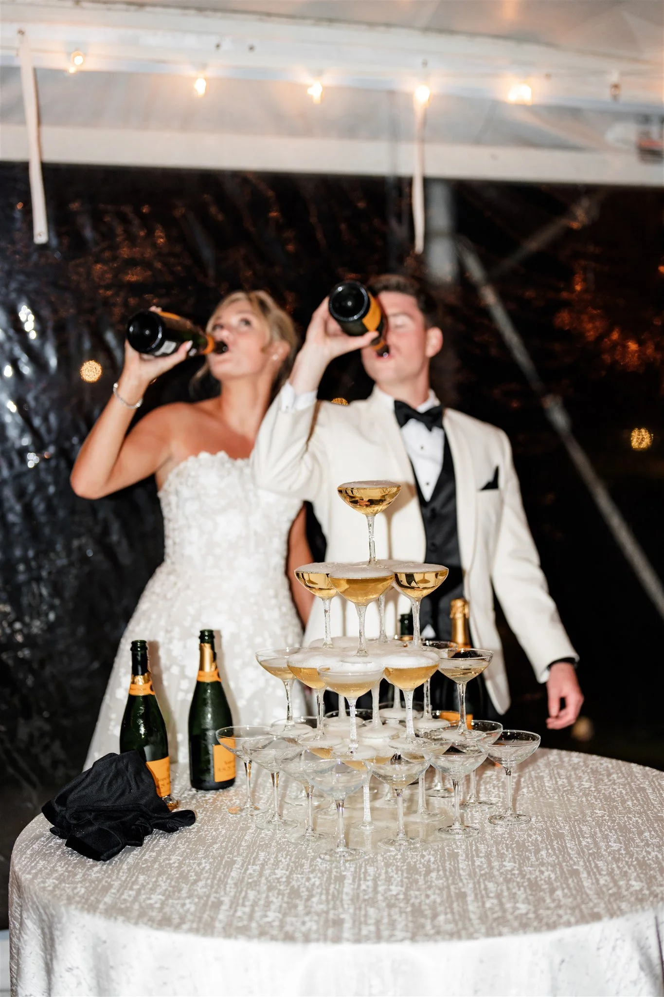 A bride and groom in formal attire celebrating with a champagne tower and toasting with bottles of champagne at a wedding reception.