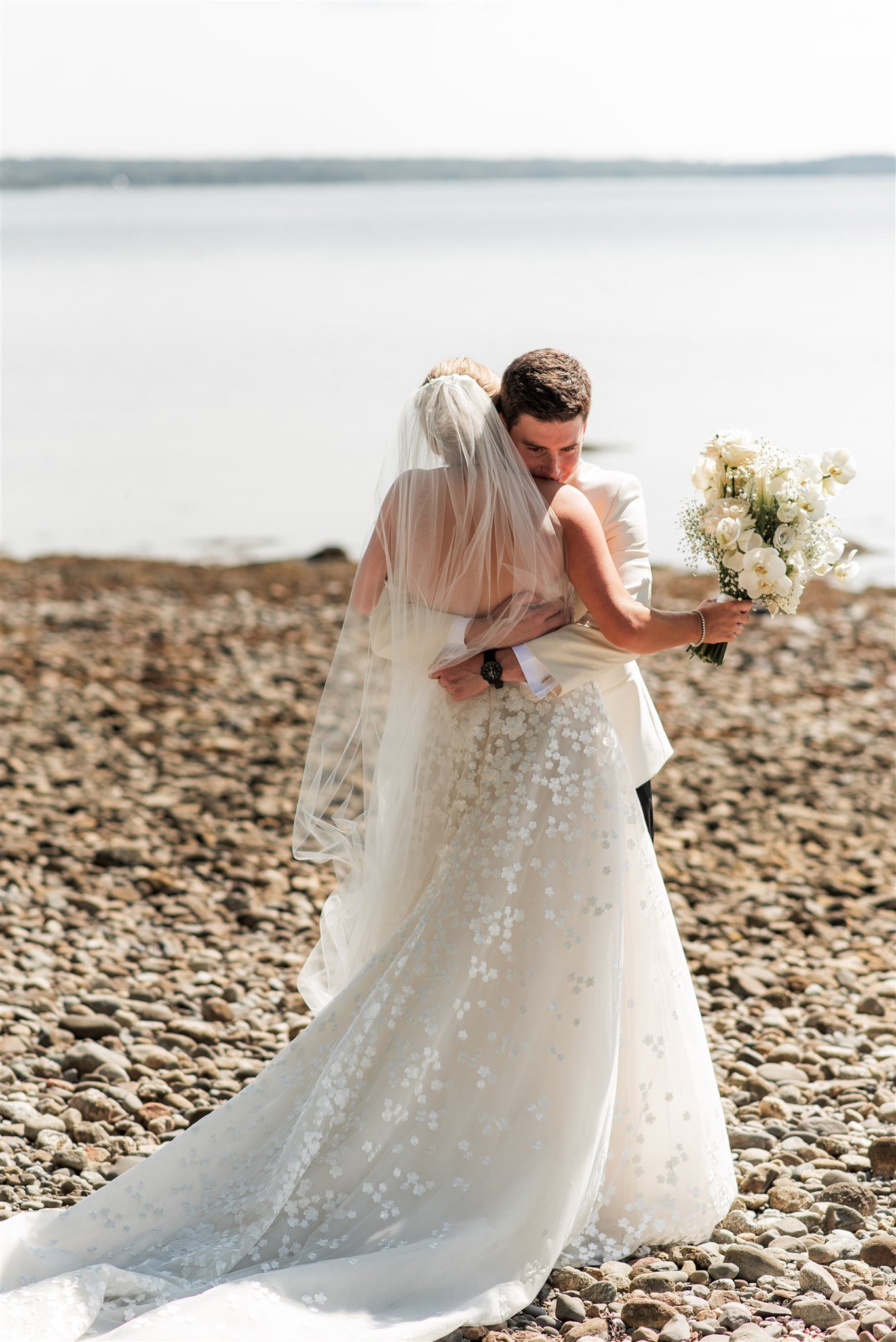 A bride and groom hugging on a rocky beach during their wedding, with the bride holding a bouquet of white flowers.
