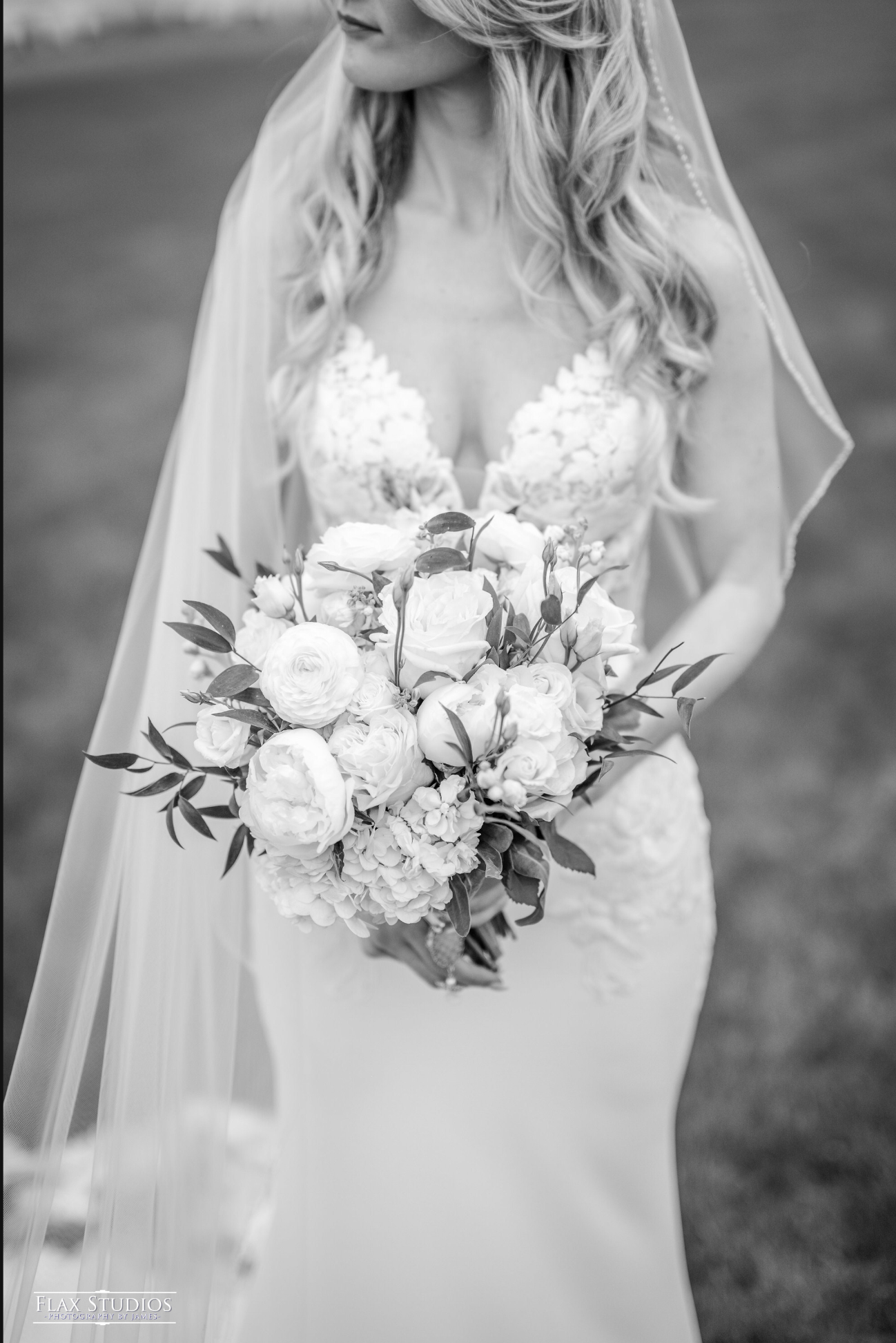 A bride in a white wedding gown is holding a large bouquet of white roses and other flowers, with her face partially visible in black and white.
