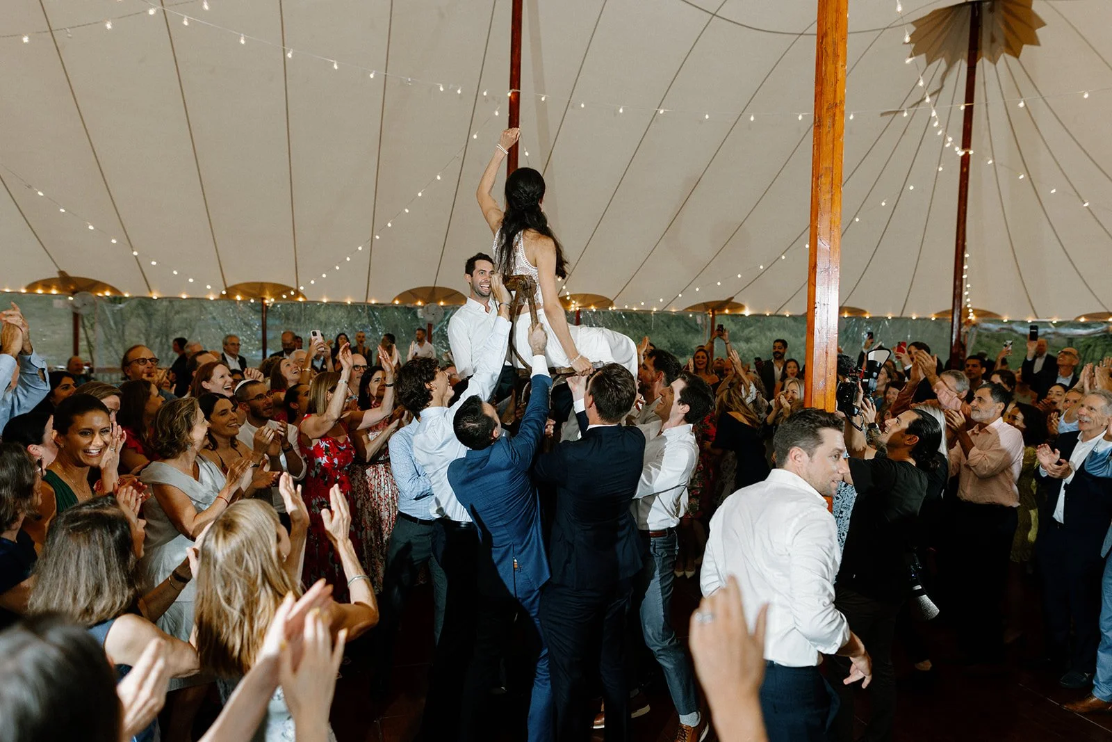 People celebrating under a large tent, with a woman sitting on a swing in the center, surrounded by a crowd clapping and cheering.