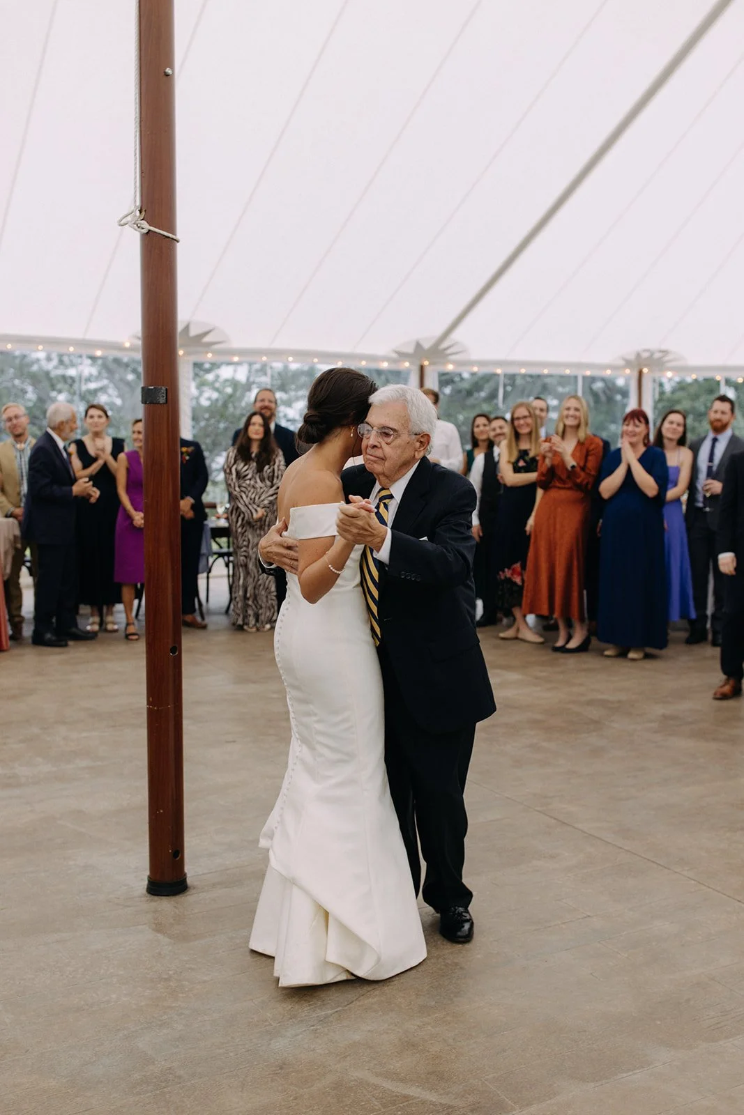 A bride and an elderly man dancing together at a wedding reception under a large tent, with guests watching and clapping in the background.