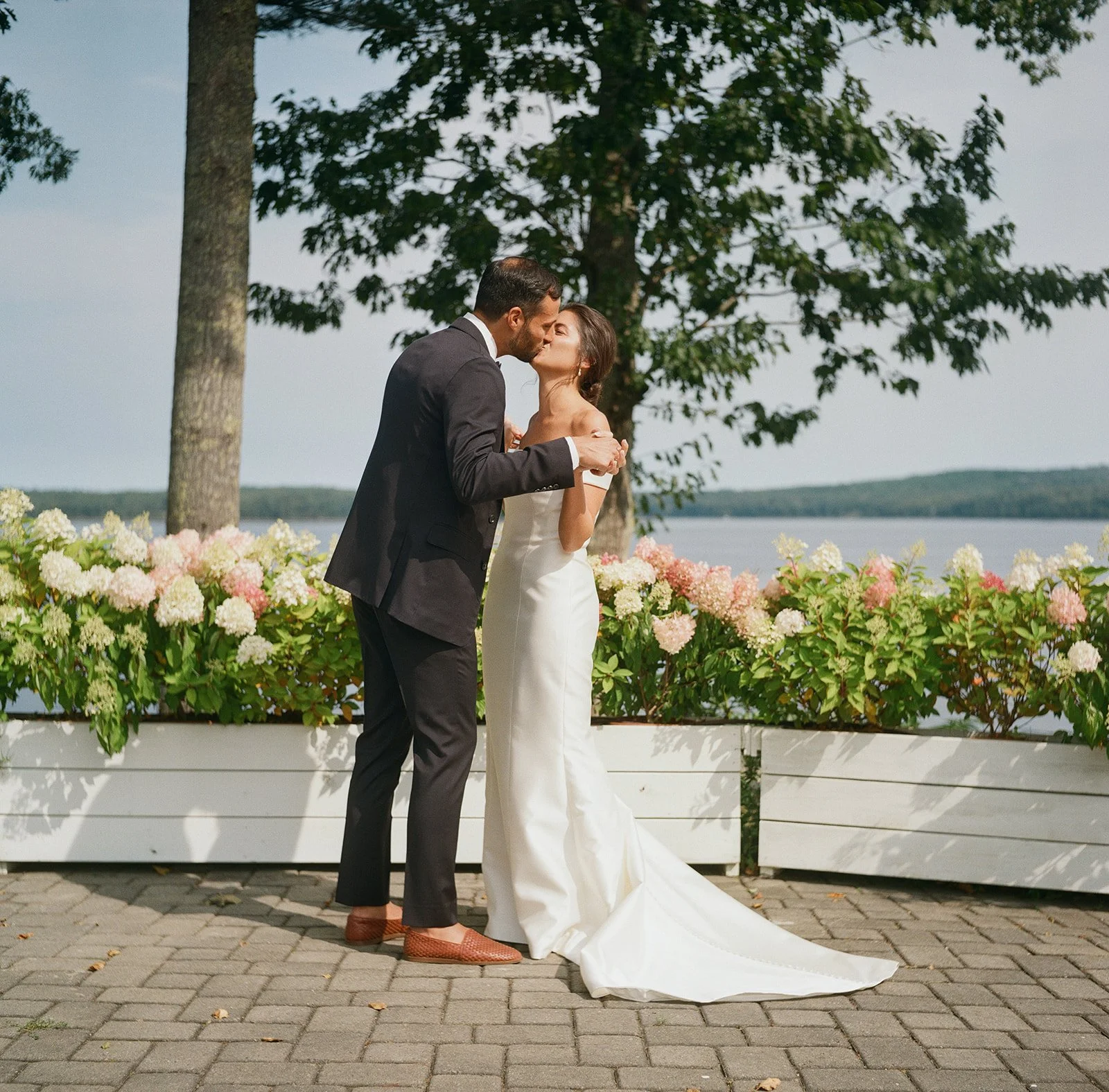 A couple in wedding attire sharing a kiss outdoors near a lake, with a row of blooming hydrangeas and green trees in the background.