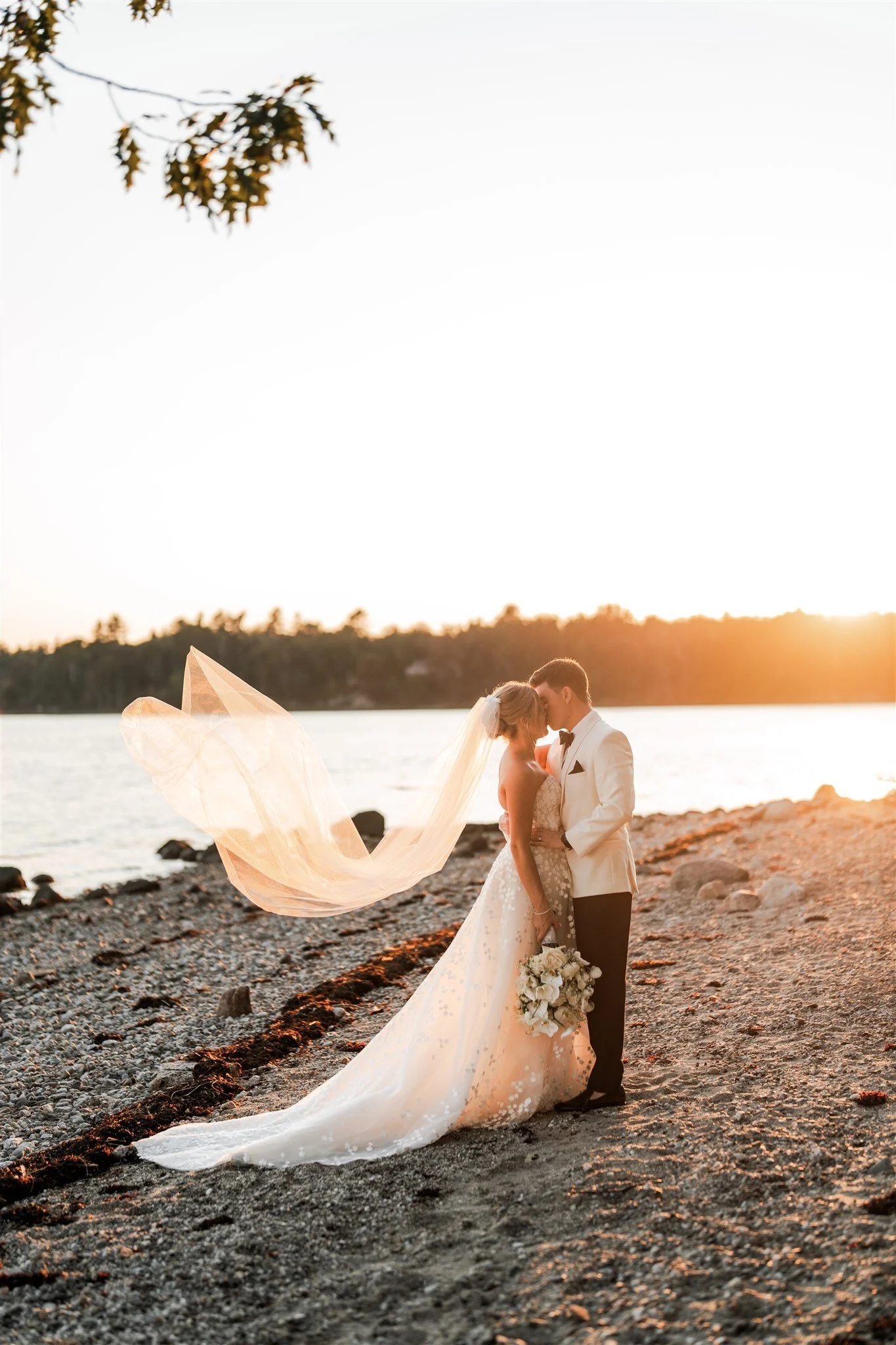 A bride and groom standing closely on a rocky beach at sunset, embracing and about to kiss, with the bride holding a bouquet of white flowers and wearing a flowing wedding dress with a long veil.
