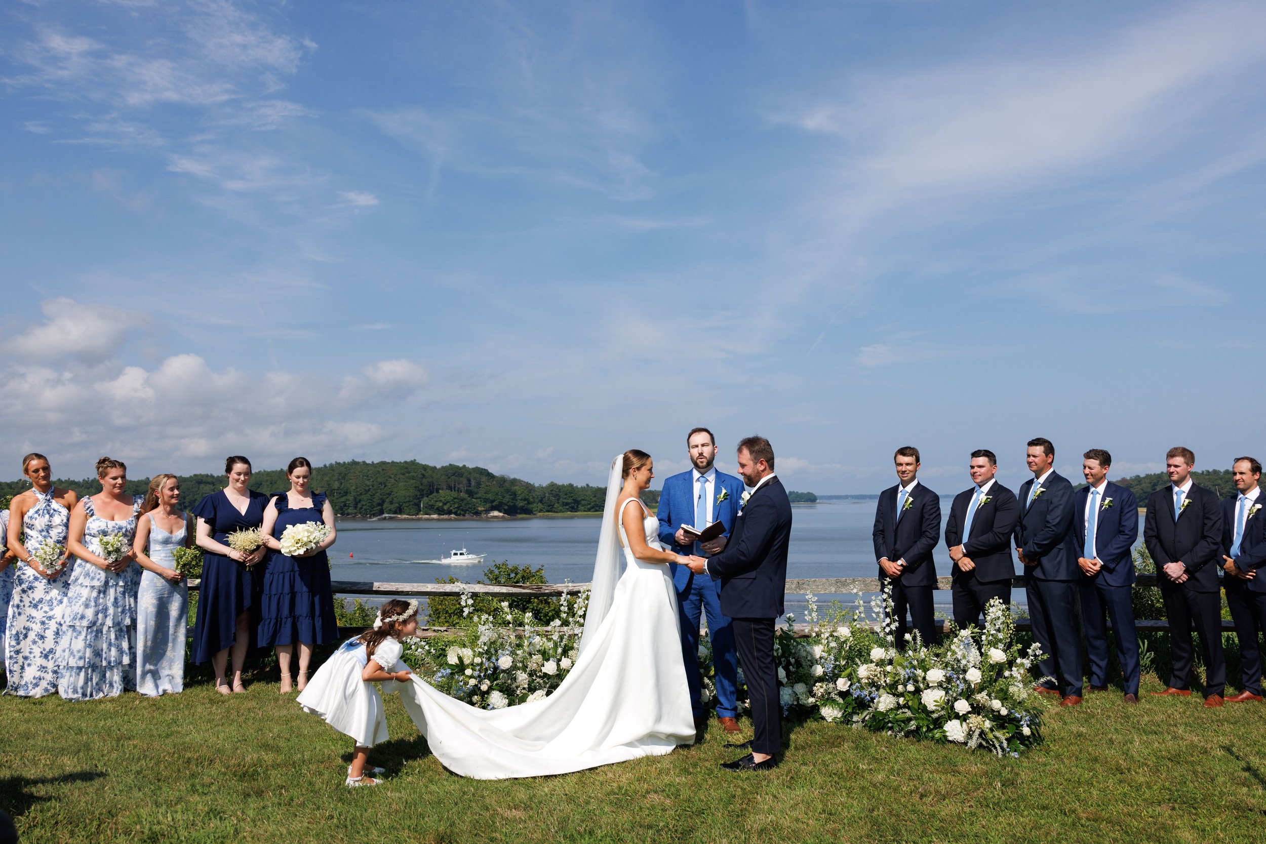 A wedding ceremony taking place outdoors near water with a bride and groom standing in front of officiant, surrounded by bridesmaids and groomsmen, overlooking a lake with boat and greenery