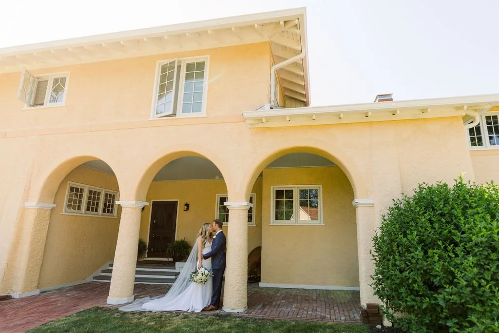 A bride and groom standing under arched porch of a yellow house, holding hands and gazing at each other, with the bride in a white wedding gown and veil, and the groom in a dark suit, surrounded by green foliage.