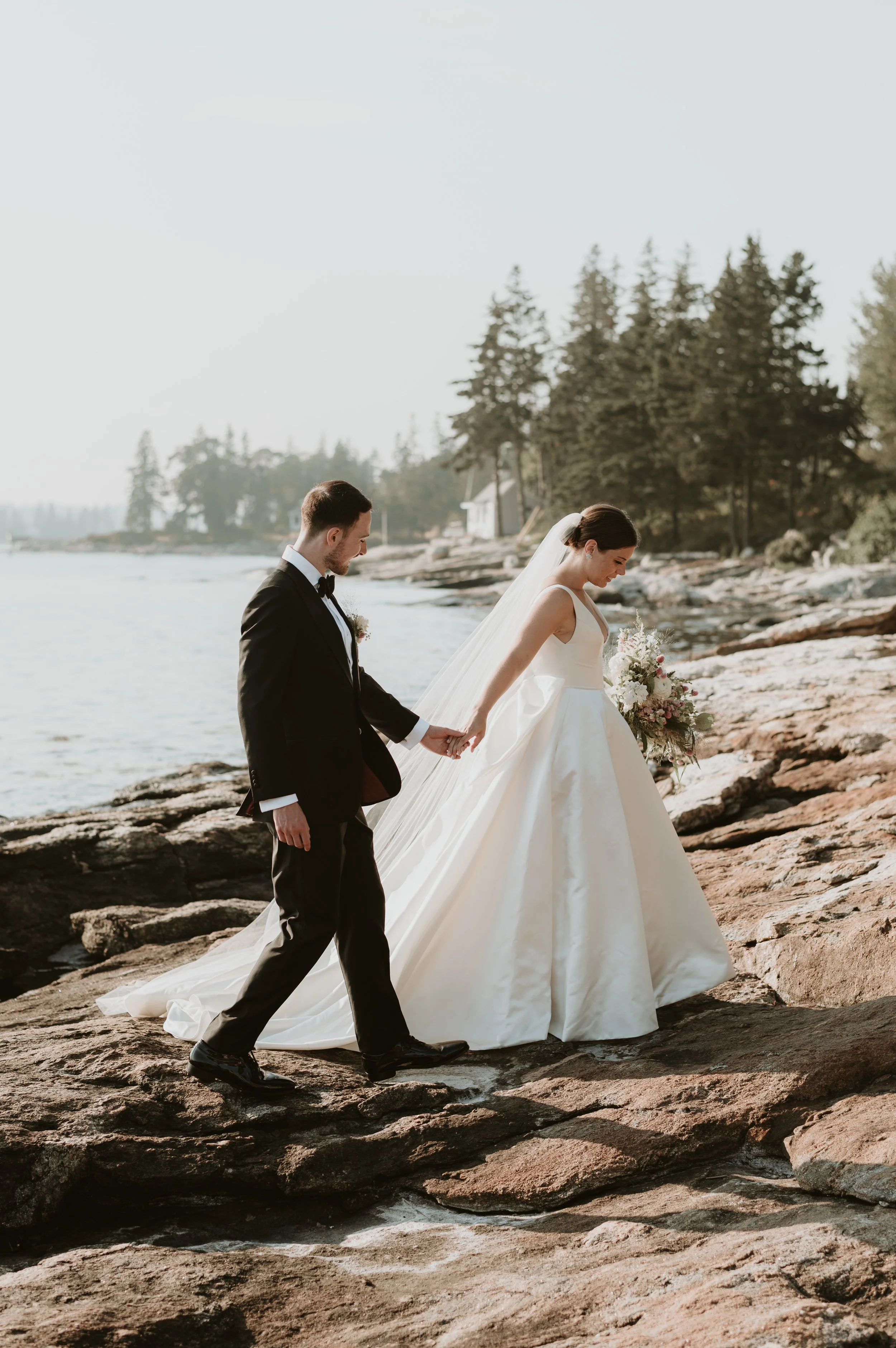Bride and groom walking hand in hand on rocky shoreline, overlooking water with trees in background, during wedding ceremony.