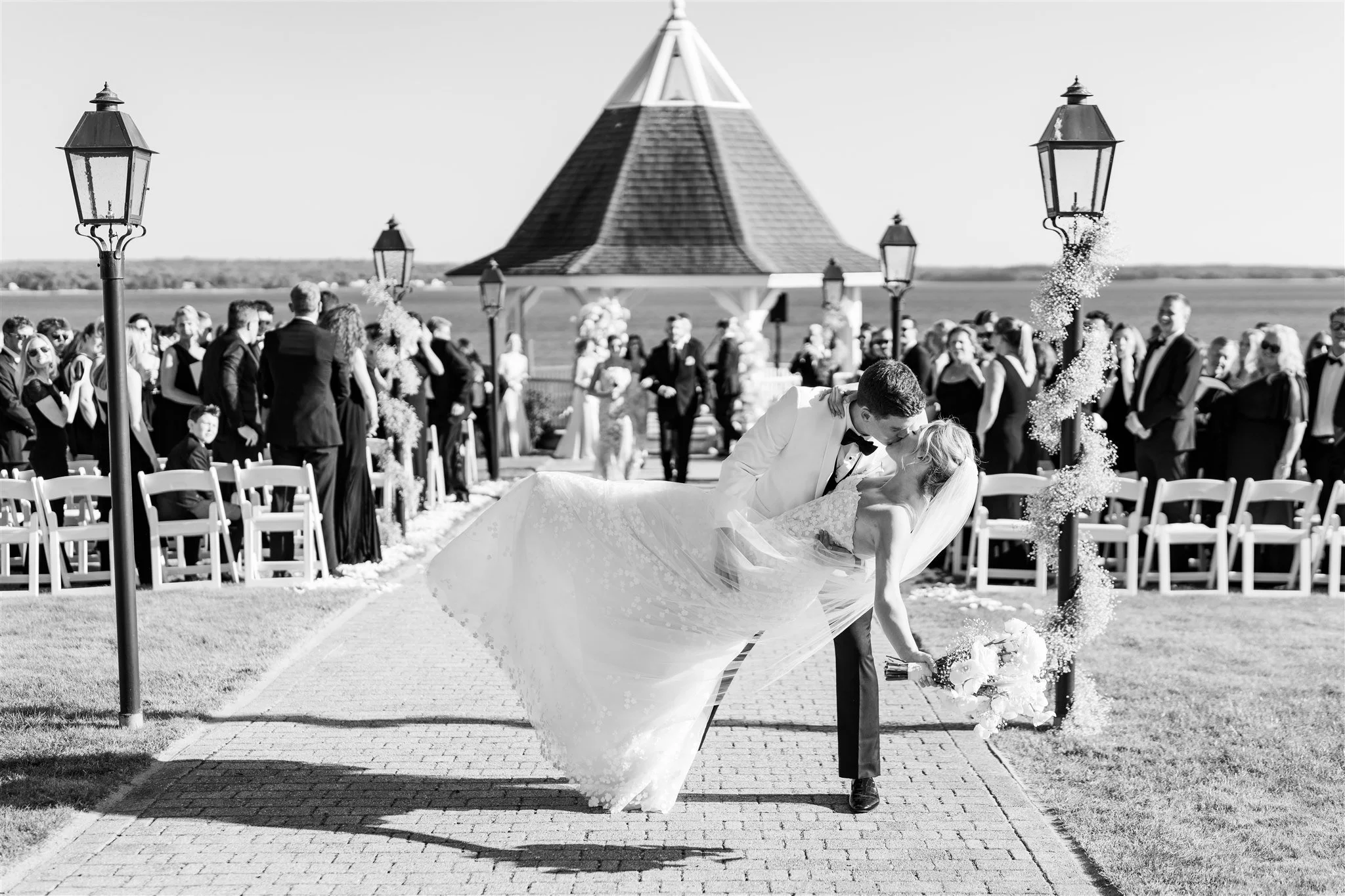 A bride and groom share a kiss during their outdoor wedding ceremony in black and white. The groom dips the bride, who holds a bouquet, with guests and a gazebo in the background.