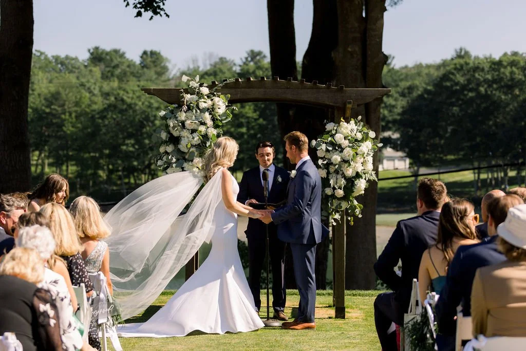 A wedding ceremony taking place outdoors under a wooden arch decorated with white flowers, with a bride and groom exchanging vows, surrounded by seated guests on a grassy area with trees in the background.
