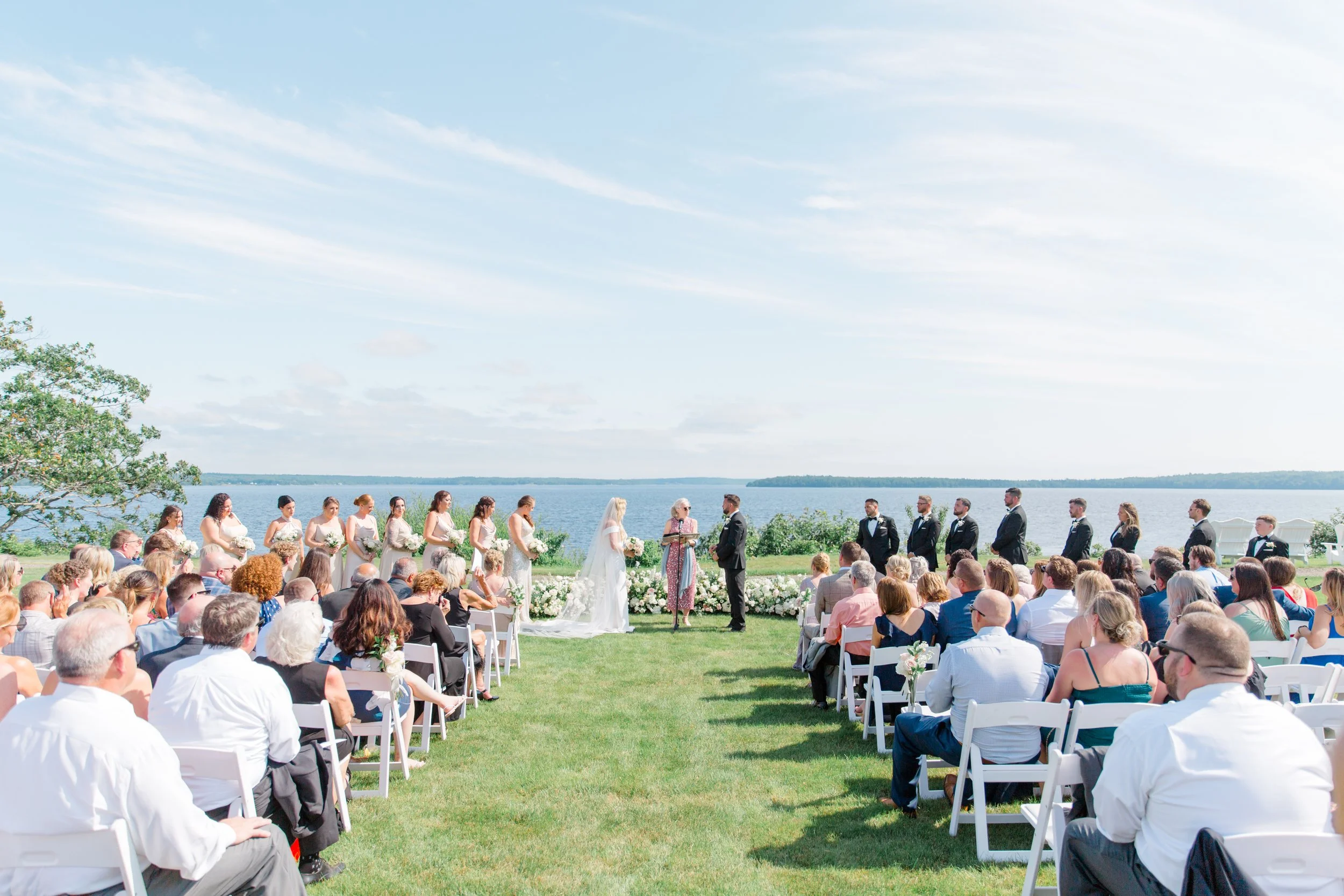 Outdoor wedding ceremony by a lake with guests seated and the bride and groom exchanging vows.