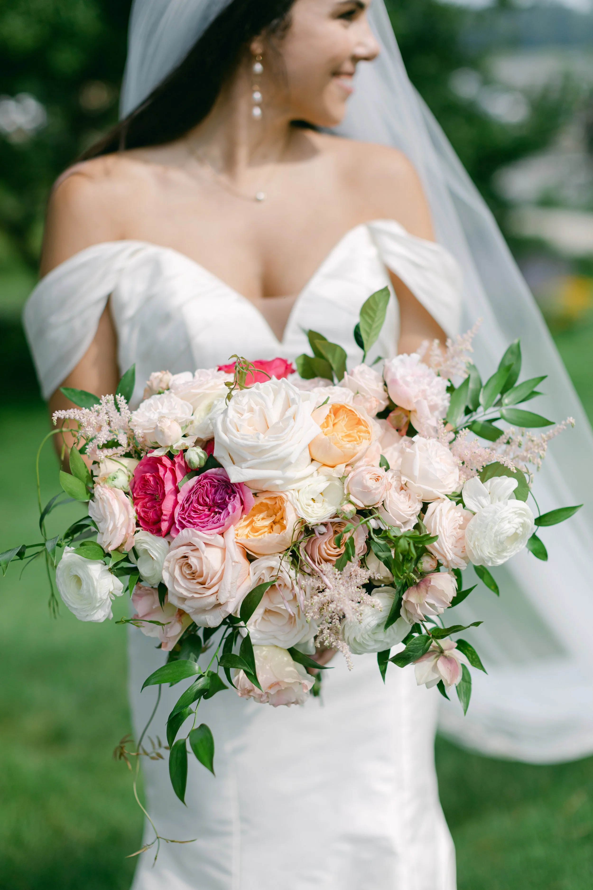 Bride holding a large bouquet of light pink, white, and peach flowers with green leaves, outdoors on a grassy area.